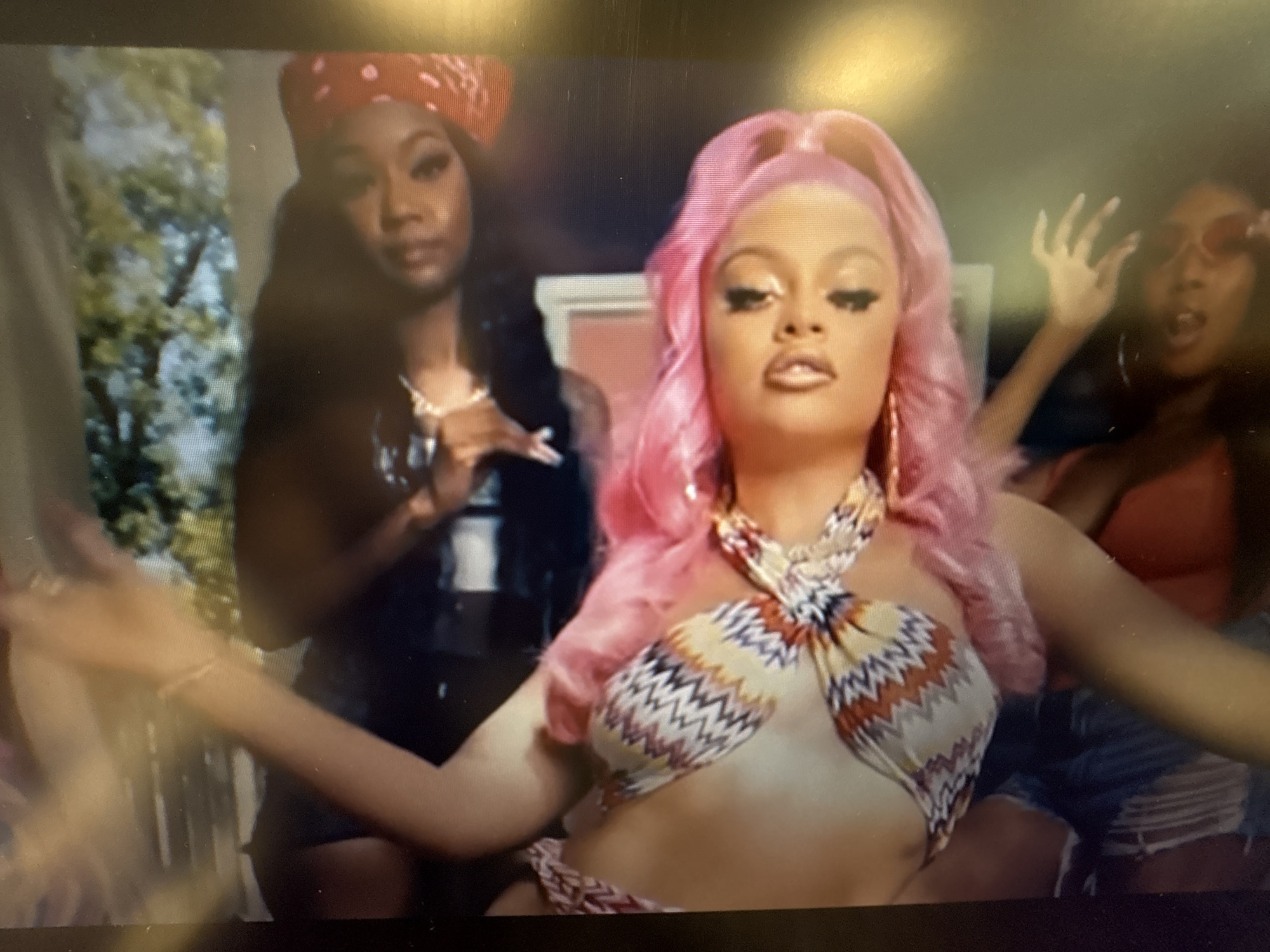 The image depicts a medium shot of three young women in an outdoor setting. The woman in the foreground, with bright pink hair styled in a high ponytail, wears a colorful, geometric-patterned bikini top and appears to be looking directly at the camera with her arms outstretched. To her left, a woman with dark hair and a red bandana is partially visible, also looking towards the camera. To the right of the foreground woman, another woman with dark hair and red-tinted sunglasses is reaching up with one hand, her mouth open as if speaking or exclaiming. The background is slightly blurred, showing greenery and what appears to be part of a building structure. There is no identifiable information in the image to suggest the location is Sofia, Bulgaria.