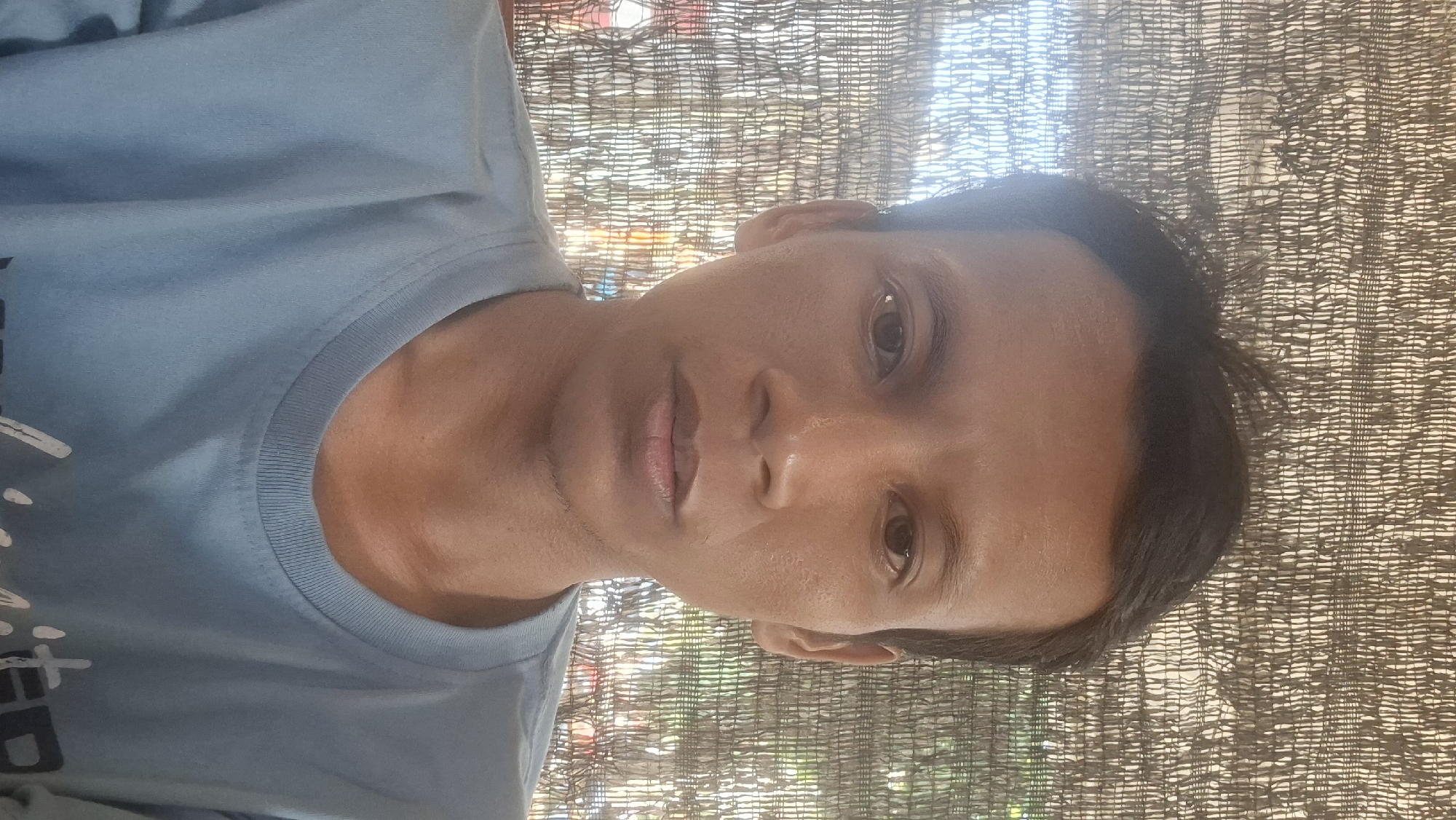 A man in a light blue t-shirt is captured in a selfie, facing forward. He has dark hair and brown eyes. Behind him, a textured, woven background of brown and white material is visible. The backdrop suggests an outdoor setting, possibly in Ngimbang, Indonesia, given the tropical foliage and light visible through the weave. The man's expression is neutral, and his posture is relaxed, with his shoulders visible as he wears the t-shirt.