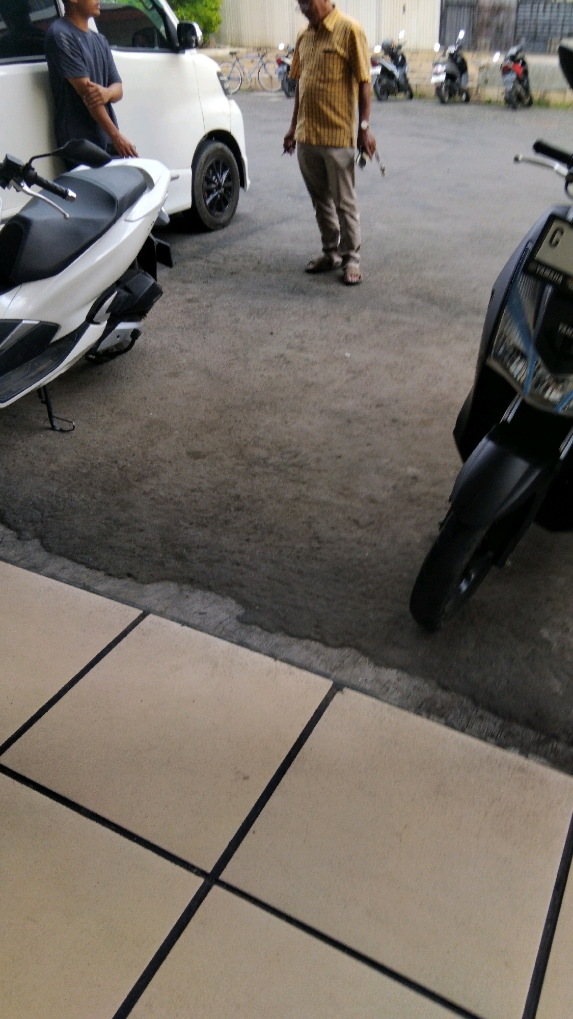 The image depicts an outdoor scene, likely in the city of Pekalongan, Indonesia, with a focus on vehicles and two individuals. In the foreground, a white scooter is parked, and to its right, a dark-colored scooter is partially visible, its front wheel and fender prominent. In the background, a white van is parked on the left side of the frame. A person wearing a dark t-shirt is leaning against the white van. To the right of the van, another person, dressed in a yellow and brown striped shirt and beige pants, stands holding a pen or small object in their right hand and what appears to be a set of keys in their left. The ground is paved with asphalt, with a tiled pathway visible in the lower left portion of the image. Several other motorcycles are parked further in the background, suggesting a public area or parking lot. The lighting indicates it is daytime.