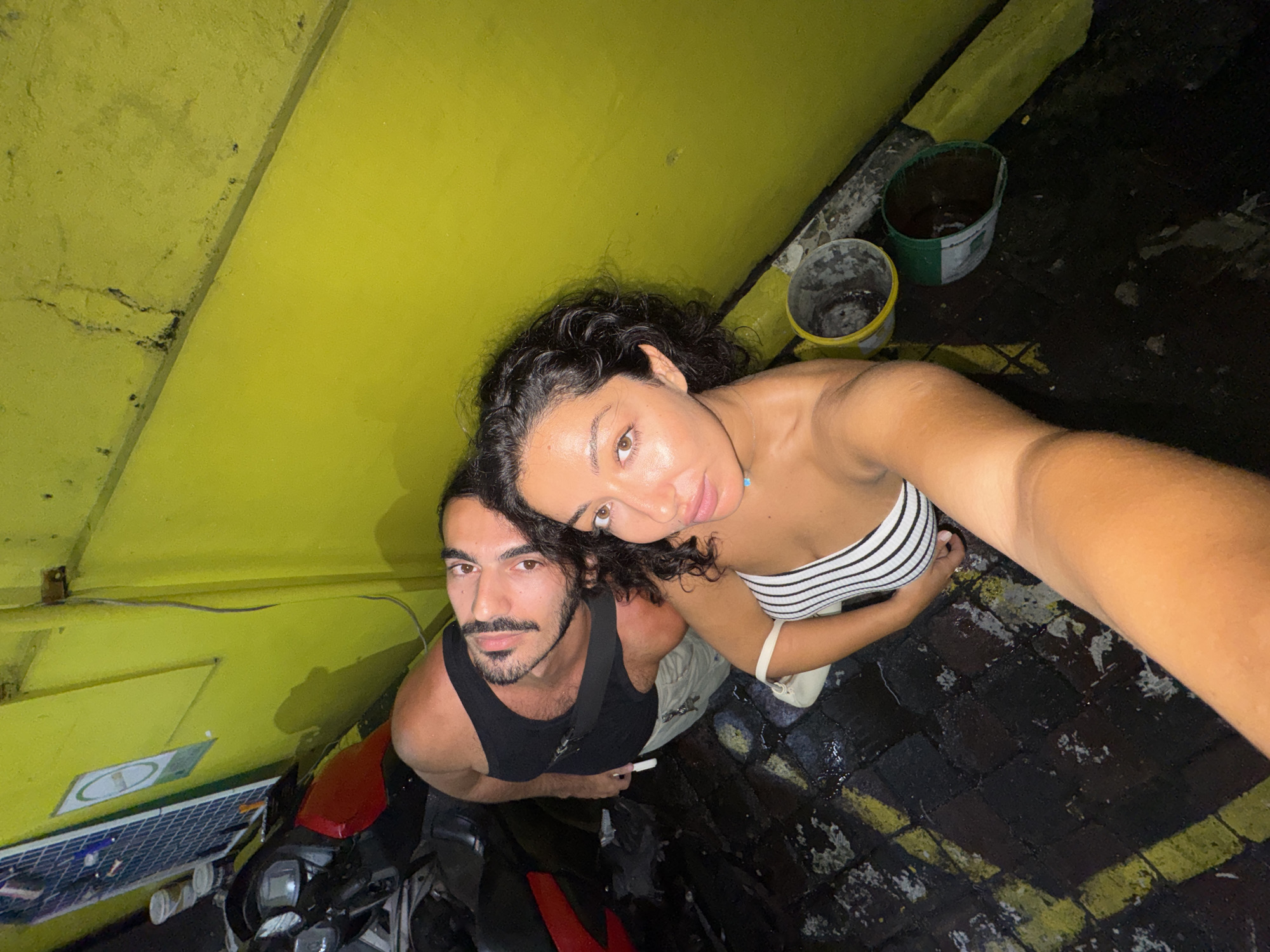 Captured from an overhead perspective, the photograph depicts a man and a woman posing for a selfie in North Kuta, Indonesia. The woman, positioned closer to the camera, wears a striped tube top. The man is to her left in a black tank top. They are standing next to a motorcycle parked adjacent to a yellow building with a solar panel affixed to it. The ground consists of black bricks with yellow stripe markings. Two paint buckets sit near the top right corner.