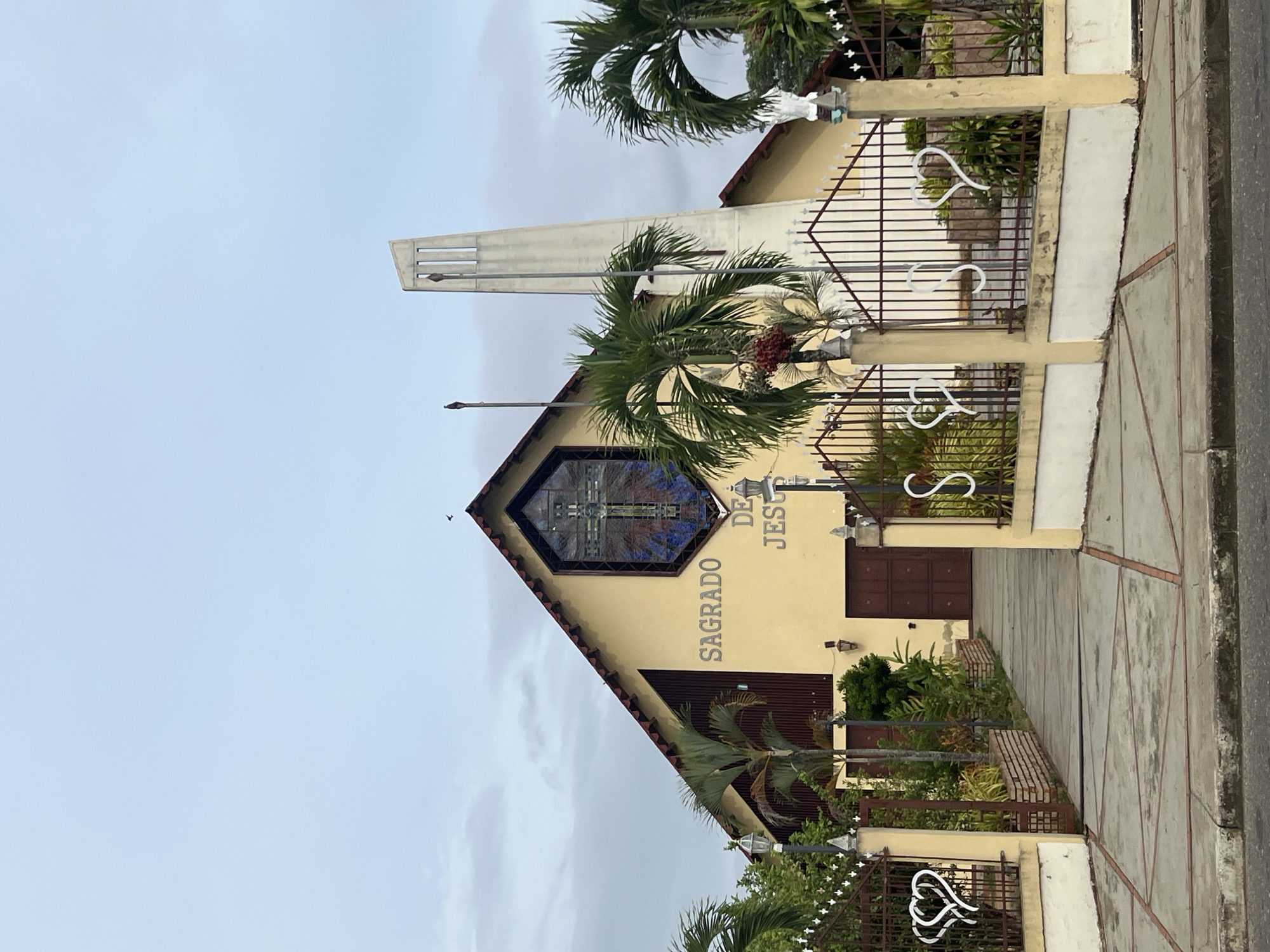 The image depicts the exterior of the Sagrado Corazon De Jesus church in Tinaquillo, Cojedes, Venezuela. The building is a light yellow color, and features a triangular-shaped front facade with a stained glass window depicting a cross. The words "Sagrado De Jesus" are written vertically on the building's front. A tall, slender steeple is visible adjacent to the church. The church is surrounded by palm trees and decorative wrought iron fencing with heart-shaped designs. The sidewalk is made of concrete. The sky is overcast.