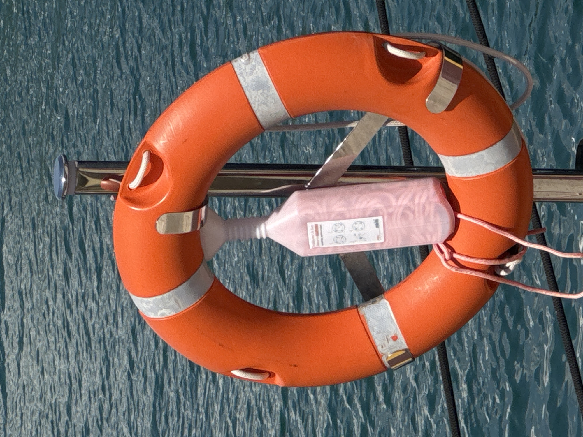 An orange life preserver is mounted vertically on a metal pole, its reflective silver bands visible. A pink plastic bottle with a white cap and a ribbed neck is suspended from the center of the life preserver. The bottle has text and symbols printed on it, suggesting it contains a liquid. The life preserver is tied with a pink rope. The background consists of rippling blue water. The scene is situated outdoors, likely near a body of water. The location is the city of Moschato, Greece.