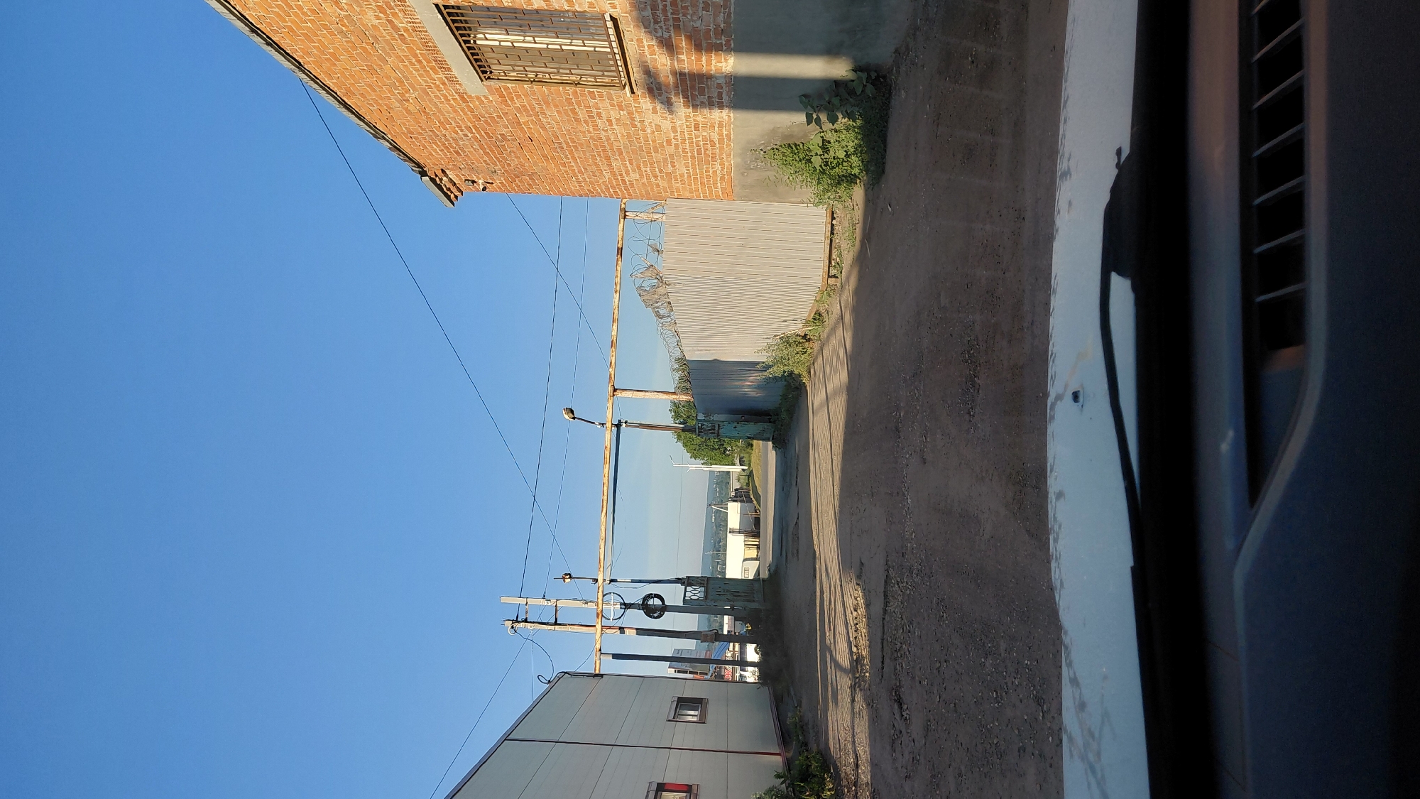 The image shows a dirt road leading through a fenced industrial area under a clear blue sky. On the left is a white building with a window. On the right is a brick building with a barred window. A metal fence with barbed wire runs along the right side of the road, separating it from the brick building. A large metal structure with lights spans the road in the middle distance. The view appears to be from inside a vehicle, with the hood and dashboard visible in the foreground. The overall impression is one of a utilitarian and somewhat neglected industrial or storage facility.