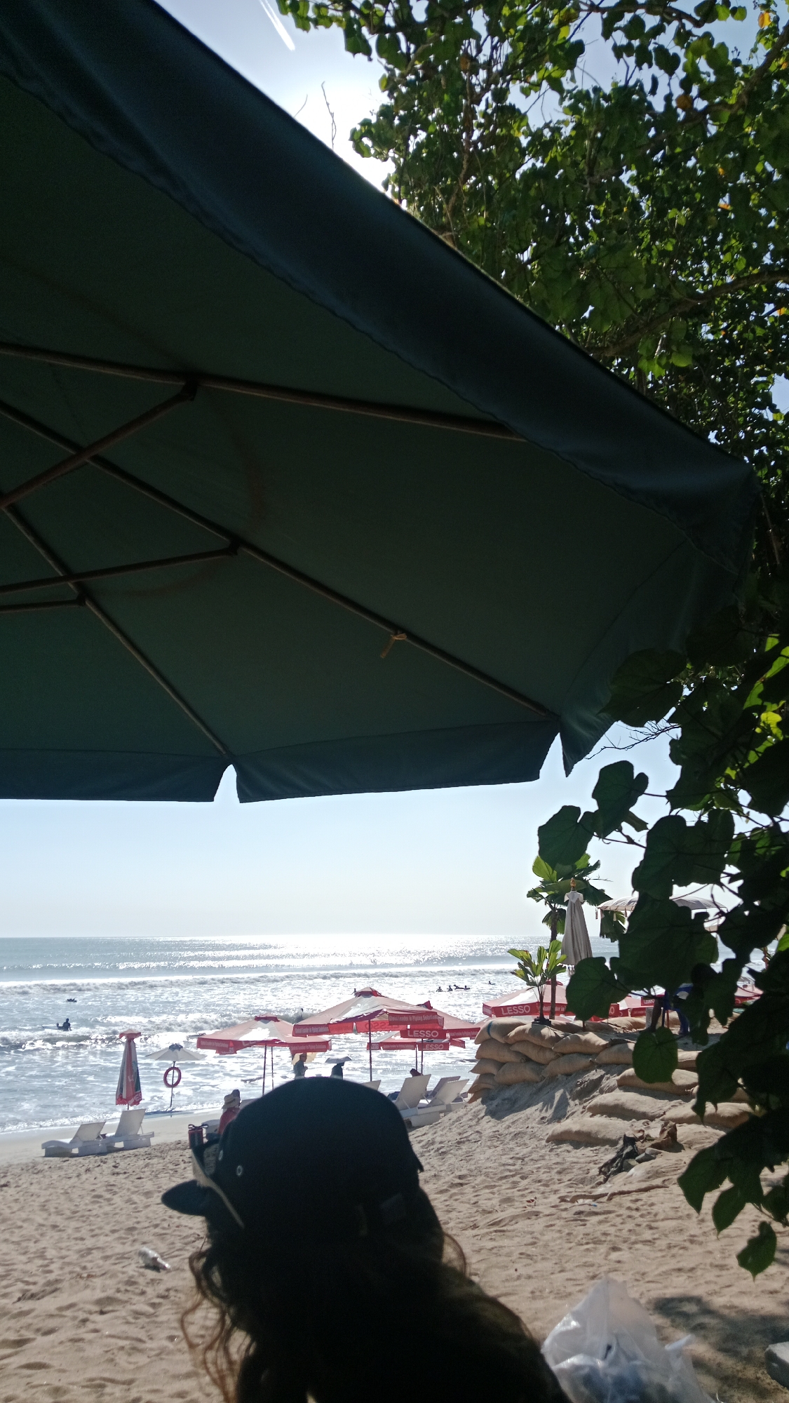 The image depicts a view from a beach umbrella, looking out towards the ocean in Kuta, Indonesia. The foreground is dominated by the underside of a large, dark green beach umbrella with visible wooden struts. Below the umbrella, a person wearing a black baseball cap and dark hair is sitting on the sand, facing away from the camera. The beach is sandy, with several white lounge chairs and red umbrellas set up in the middle ground. Beyond the chairs, the ocean is visible, with the sunlight glinting off the water's surface. Small waves are breaking near the shore. A few people are in the water, and a sandbag barrier is visible on the right side of the image, supporting some palm trees. The sky is bright and clear with a faint contrail visible. The overall scene suggests a sunny day at the beach.