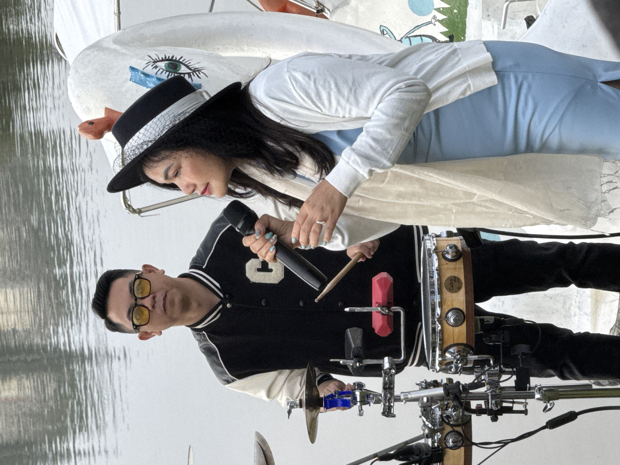 In Da Lat, Vietnam, a man and a woman are performing by the water. The man, wearing yellow-tinted sunglasses and a black and white letterman jacket, stands behind a drum set. The woman, sporting a black hat with a veil and a white cardigan over a light blue dress, holds a microphone. A large, white swan-shaped decoration is visible behind them. The water's surface reflects the overcast sky, creating a muted backdrop for the performance.