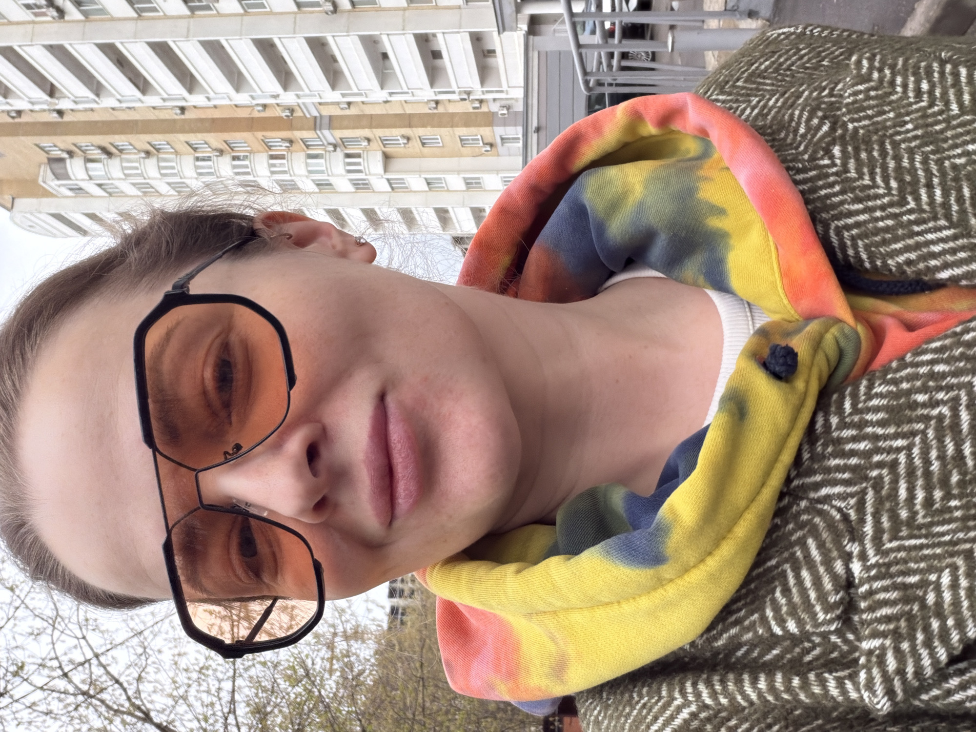 A woman wearing sunglasses and a colorful hooded sweatshirt over a white top is the primary subject. She is also wearing a herringbone-patterned coat. In the background, there is a multi-story building with numerous windows, suggestive of residential apartments, and trees with bare branches. The overall scene is a city setting. The location is likely Moscow, Russia.
