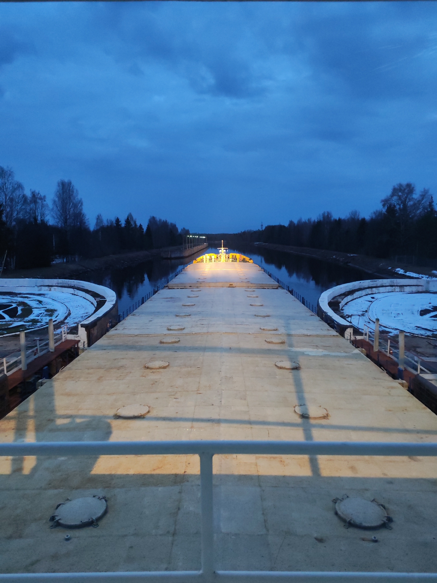 The image depicts a canal lock in what appears to be a forested area, possibly near dusk or dawn. The foreground features a concrete surface with evenly spaced circular hatches and a low white railing. To the left and right are semi-circular areas, partially covered in snow. Beyond this, the canal stretches into the distance, reflecting the ambient light. Trees line the canal banks on both sides. In the distance, lights illuminate what may be another section of the lock or a control area. The sky is a dark blue, hinting at either early morning or late evening.