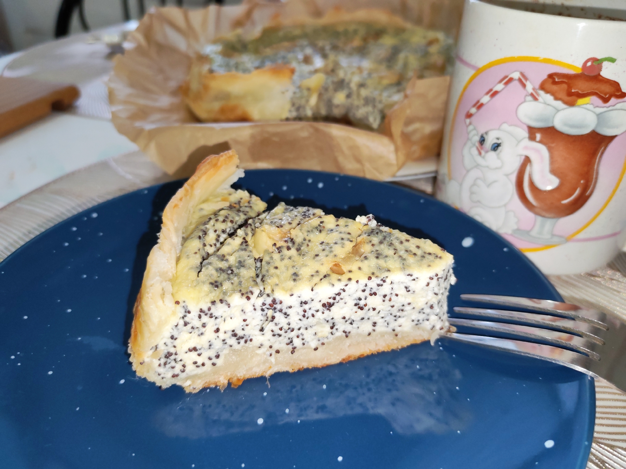 In Izhevsk, Russia, a slice of poppy seed cake sits on a dark blue plate speckled with white dots. A silver fork rests on the right side of the plate, its tines touching the cake. In the background, another piece of the cake is visible, partially wrapped in brown paper. To the right of the cake, a white ceramic mug with a pink border features a drawing of a cartoon bunny and a chocolate milkshake with a cherry on top. The table surface appears to be covered with a light-colored tablecloth.