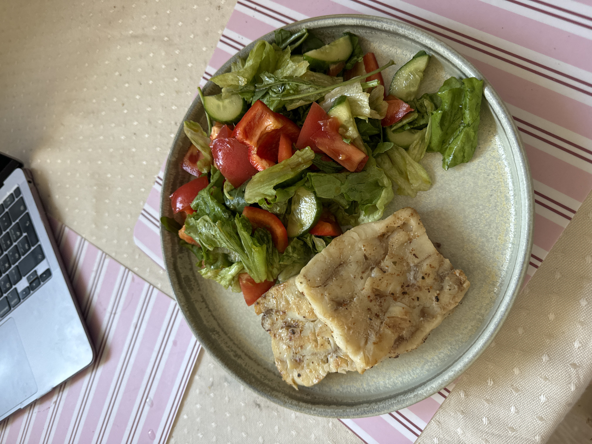 A top-down shot shows a plate of food next to a laptop. The plate holds two pieces of cooked fish and a salad with lettuce, cucumber, and red bell pepper. The plate sits on a pink and white striped placemat which is on top of a textured tablecloth. Part of a laptop is visible to the left of the placemat. The environment appears to be indoors. The location and people present are not identifiable.