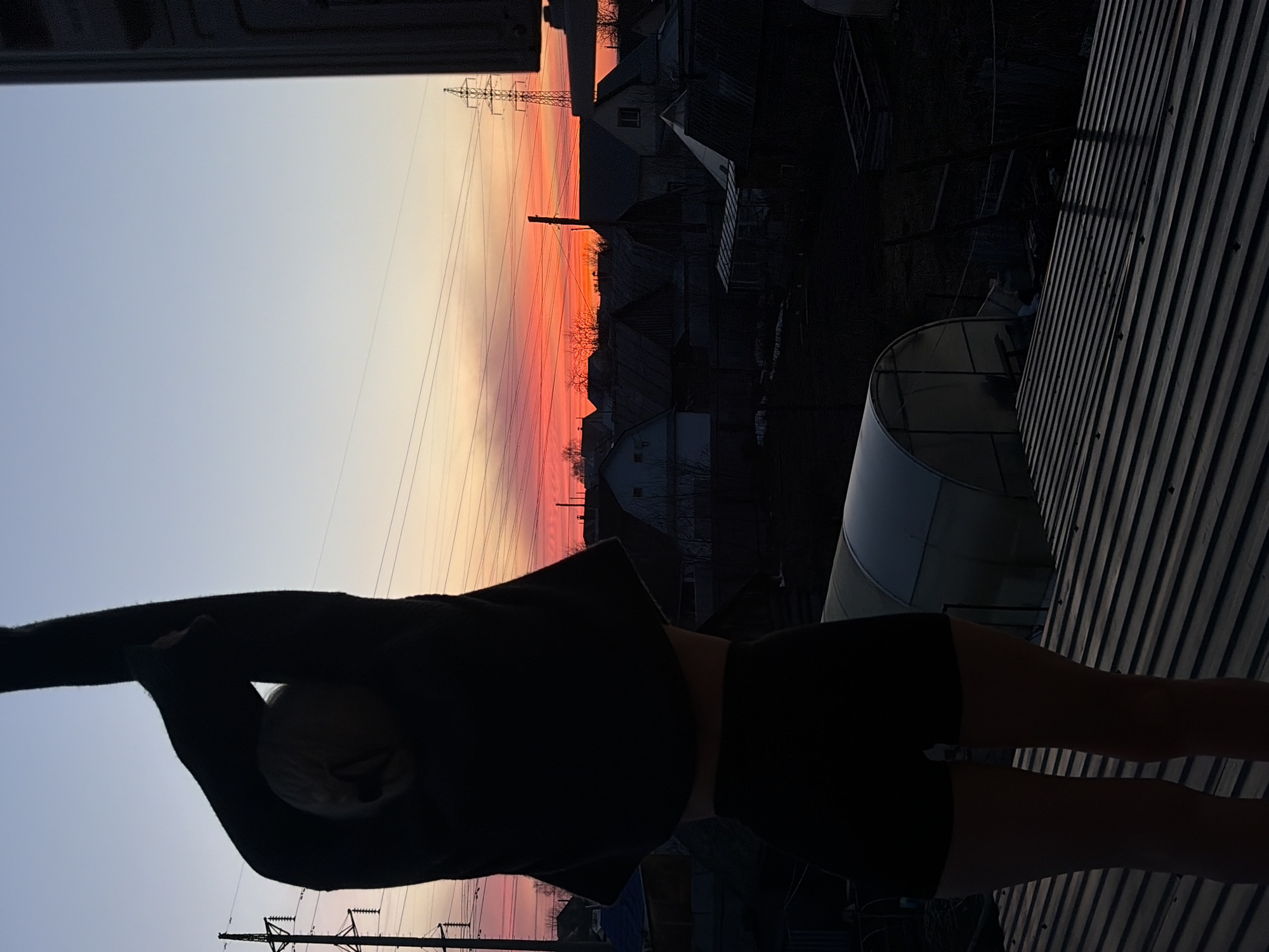 The image shows a partially obscured view of Chaikovsky, Russia, at dusk or dawn. A person wearing dark clothing is in the foreground, silhouetted against the sky. In the background are buildings, trees without leaves, and an electrical transmission tower. The sky exhibits an orange and pink gradient. Corrugated metal and a greenhouse are visible in the midground.