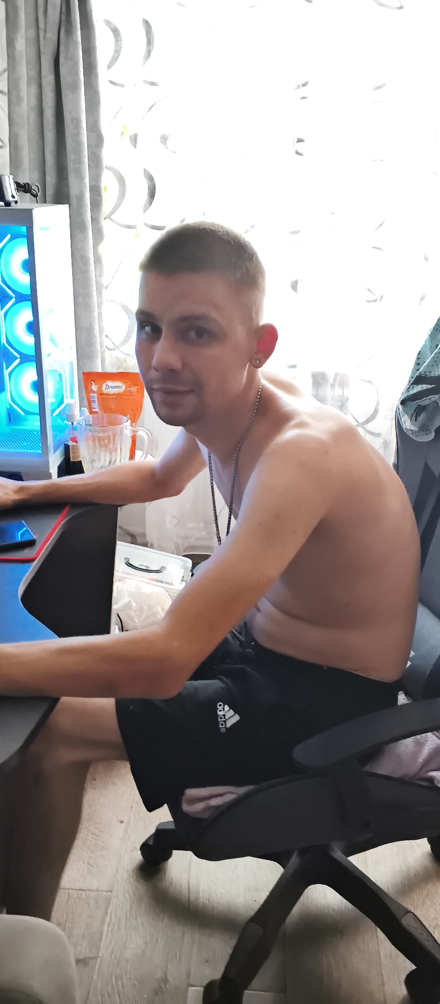 The image depicts a shirtless young man sitting in a black office chair in what appears to be a room with wood-laminate flooring and light-colored curtains. He has short, light brown hair and is wearing black athletic shorts with a white Adidas logo. A silver chain necklace is visible around his neck, and he has a gold earring in his left ear. The man is looking towards the viewer with a neutral expression. To his left, a computer tower with blue-lit fans is visible, along with a desk and a glass mug containing a beverage. An orange packet of "Dreamers" snacks is also on the desk. The overall setting suggests a casual indoor environment, possibly a home office or gaming setup, in Kondrovo, Russia.