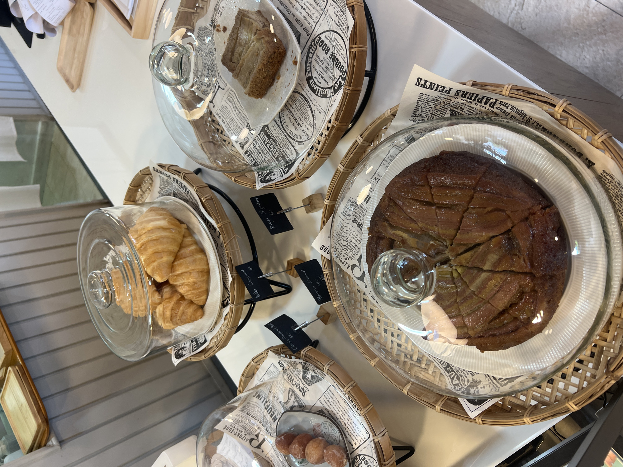 Here is a detailed description of the image:
**Scene:** An overhead shot of a display of baked goods, possibly in a bakery or cafe setting.
**Objects:**
* **Baked Goods:** There are four separate displays, each under a glass dome. These include:
* Croissants
* Doughnut balls
* A piece of banana bread
* A round banana caramel cake cut into slices.
* **Display Elements:** The baked goods are placed in round wicker baskets. Each basket has a piece of paper with newspaper print.
* **Price Tags:** Small black tags with white writing and prices are displayed next to each item. They appear to indicate the names and prices of the pastries, such as "Banana Caramel - 45.-" and "Classic Doughnut Ball 35.-".
**Environment:**
* The display is on a clean, white countertop. The lighting is bright and appears to be natural.
* A wooden object can be seen at the edge of the shot and could be part of the counter or more displays.
**Possible location:**
* The city of Unknown, Unknown. It is impossible to define a location with the details included in the image.