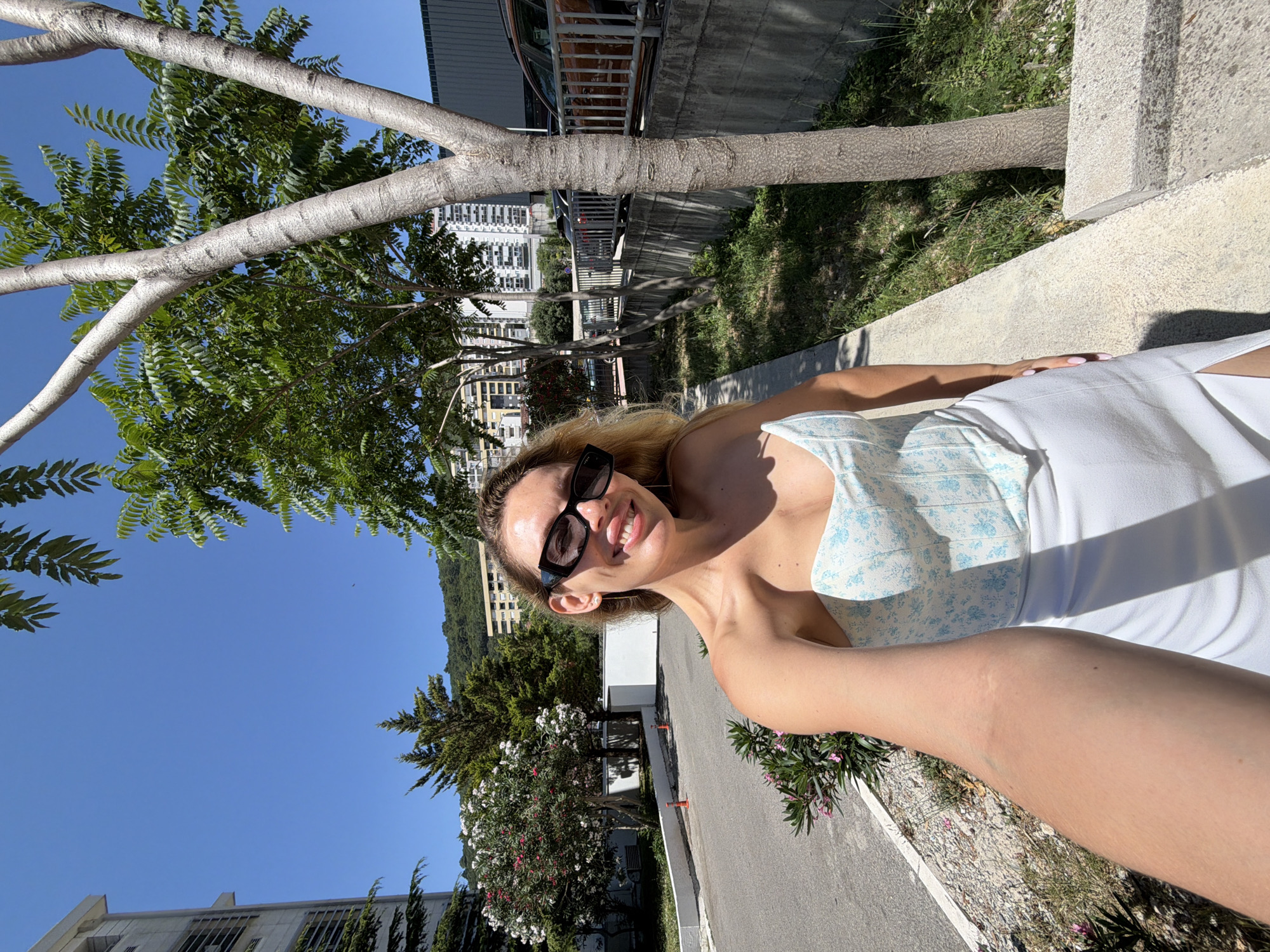 A woman with blonde hair and sunglasses takes a selfie outdoors. She is wearing a strapless corset top with a blue floral pattern and a white skirt. She is standing on a concrete path next to a tree, with buildings and hills in the background. The scene appears to be in Budva, Montenegro, based on the architectural style of the buildings and the hilly terrain. The weather is sunny with a clear blue sky.