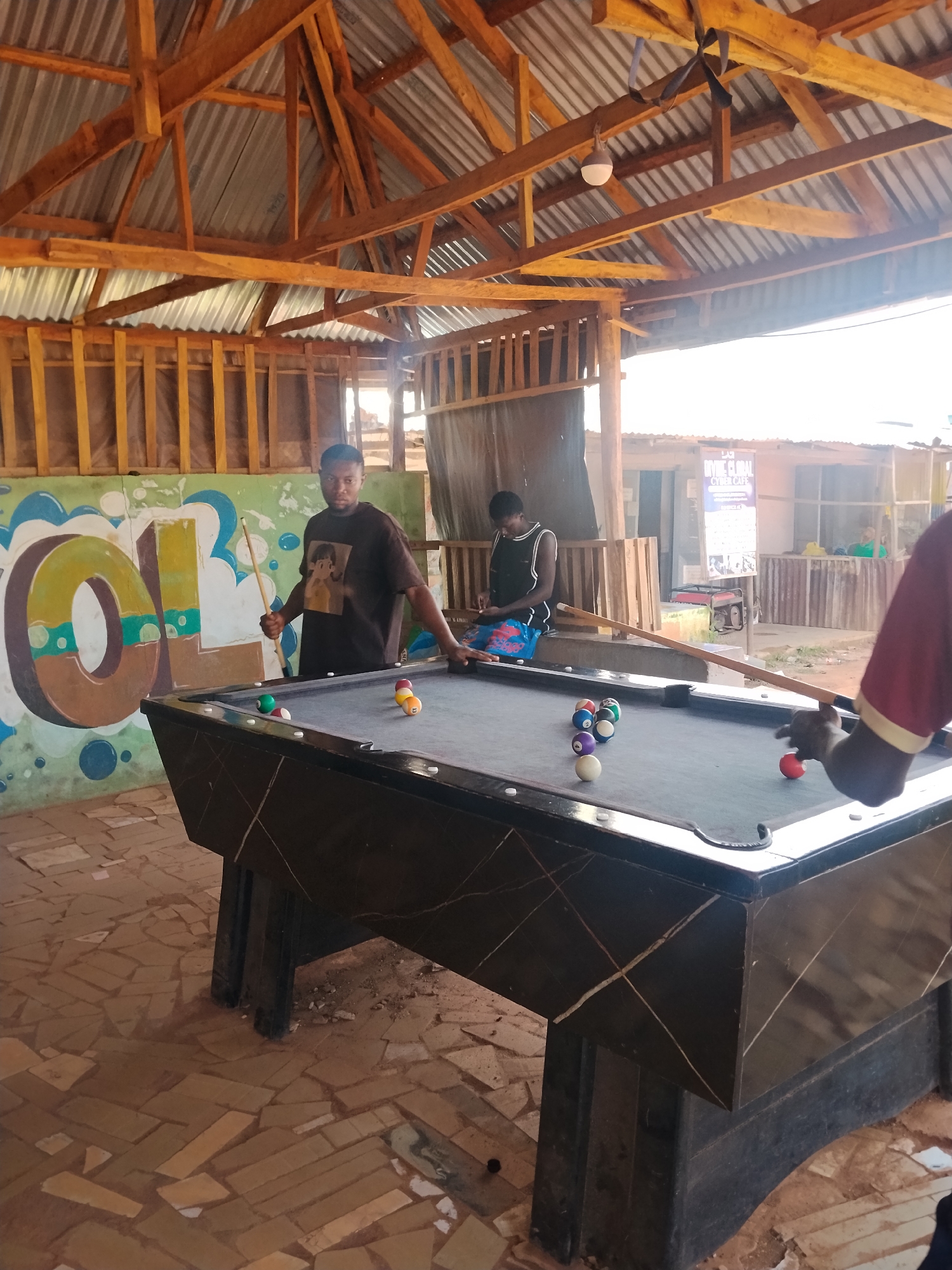 In Durumi, Nigeria, an outdoor covered area with a corrugated metal roof supported by wooden beams houses a billiards table. Three individuals are present. One man, wearing a dark t-shirt with a cartoon image, stands at the table holding a cue stick, appearing to prepare for a shot. Another person, dressed in a black tank top and blue shorts, sits on a wooden bench to the left of the table, looking down. A third person, partially visible from the shoulder down, is in the foreground on the right, also near the table. Several billiard balls are scattered on the table. A colorful mural, featuring large block letters and abstract shapes, adorns the wall behind the billiards table. The ground is paved with irregular, earthy-toned tiles. In the background, buildings with corrugated metal roofs and wooden structures are visible, suggesting a semi-developed or informal urban setting.