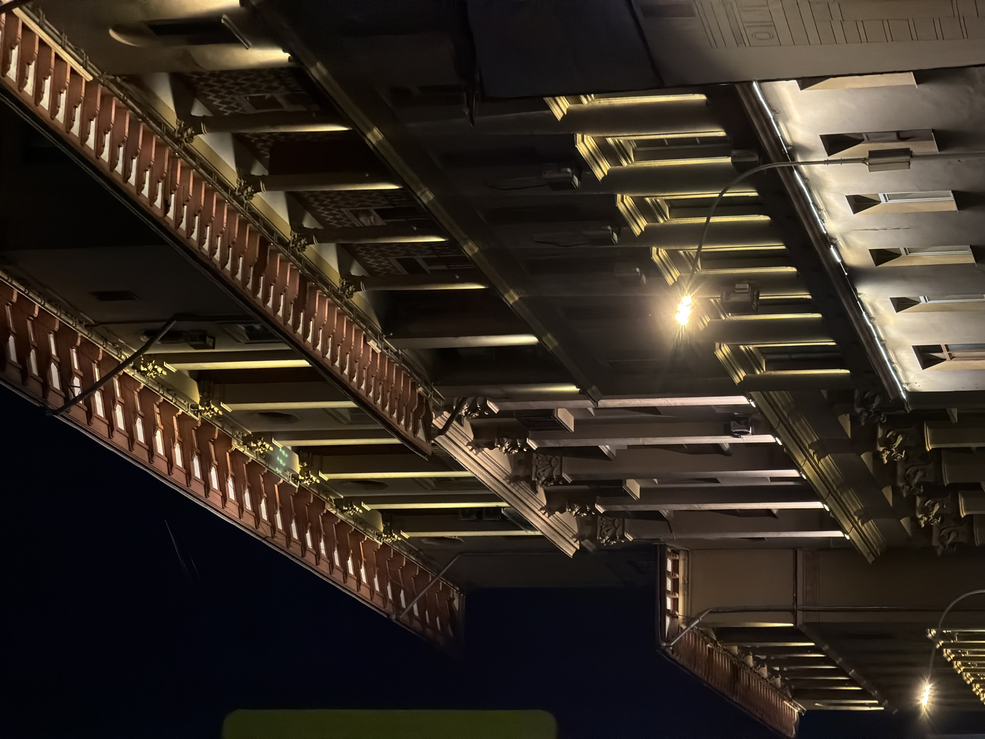 The image shows the underside of a bridge at night. The structure features a complex system of beams, supports, and decorative elements. A light fixture is visible, providing illumination. The location is the Luzhniki Metro Bridge in Moscow, Russia.