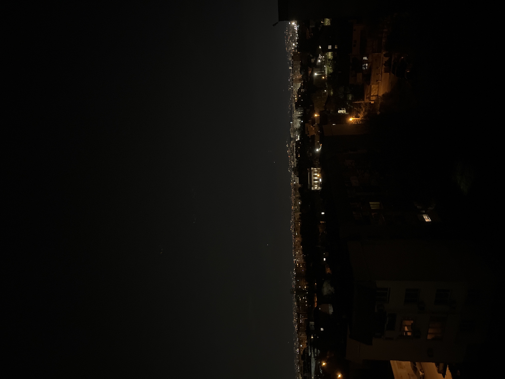 The image presents a nighttime view of what appears to be an urban landscape. The scene is dominated by darkness, with only sparse light sources illuminating the environment. Based on the layout and overall details, the location is likely Sofia, Bulgaria. The right side of the frame showcases buildings and structures with scattered lighting, suggesting residential or commercial areas. The left side shows a dark and vast area, probably the night sky or a distant dark part of the city. There is no immediate indication of people, specific subjects, or actions. The environment appears to be a typical urban setup at night, with minimal ambient illumination, and distant city lights.