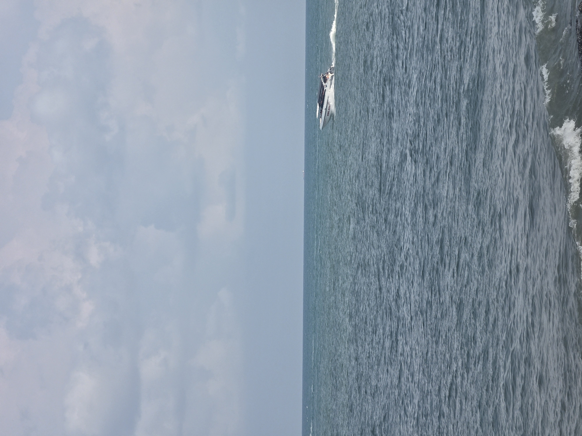 The image depicts a seascape at Pattaya City, Thailand. The left portion is the sky with scattered clouds, while the right portion features the ocean. A speedboat is traversing the water, leaving a trail of wake behind it. The shore is visible on the far right edge of the image, indicated by the presence of wave action.