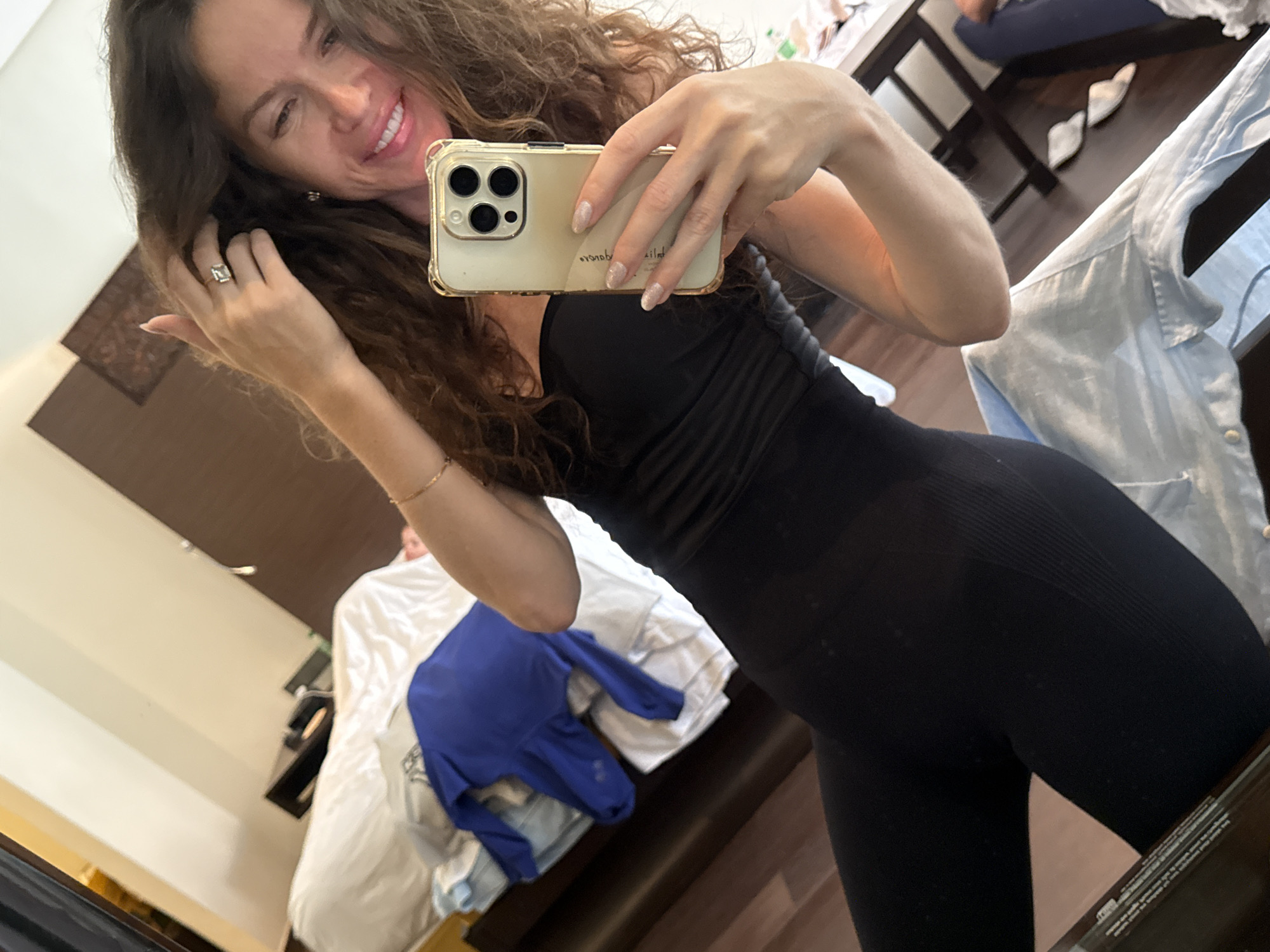 A woman in a black strapless top and black leggings is taking a mirror selfie in a bedroom. She has long, curly brown hair and is wearing a ring on her right index finger. A phone with a gold case is held in her right hand. In the background, a bed is partially visible with white and blue linens. There is also a wooden dresser and a glimpse of a table with chairs. The lighting suggests it is daytime.