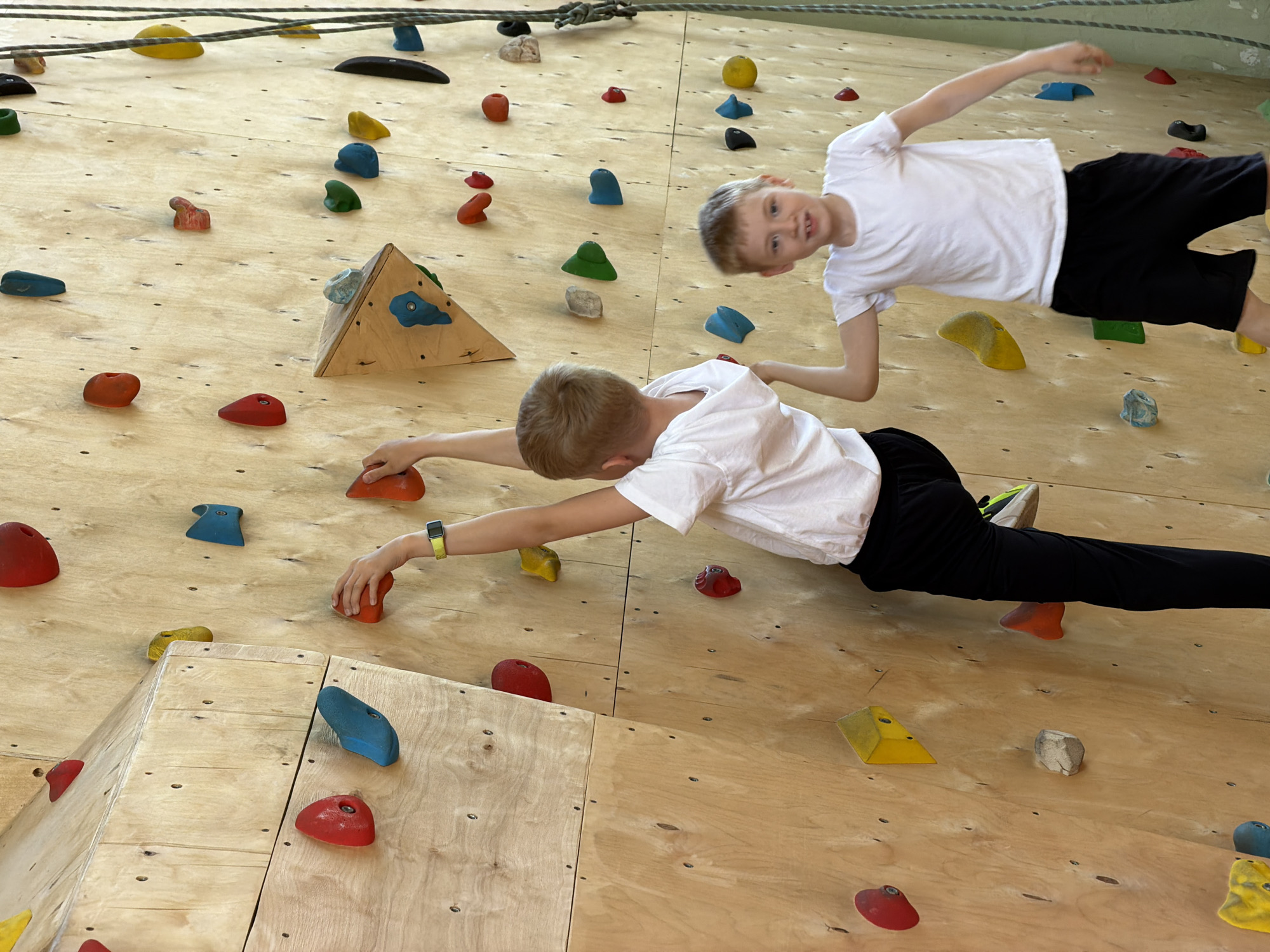Two children are climbing an indoor climbing wall. The wall is made of light-colored wood and has numerous colorful climbing holds attached to it. One child is positioned lower on the wall, reaching for a hold with his right hand. He is wearing a white shirt, black pants, and a watch on his left wrist. The other child is positioned higher on the wall, with his body turned towards the viewer. He is also wearing a white shirt and black pants. The background of the image includes more of the climbing wall and the ceiling. The location is likely an indoor recreational facility. The city is Izhevsk, Russia.
