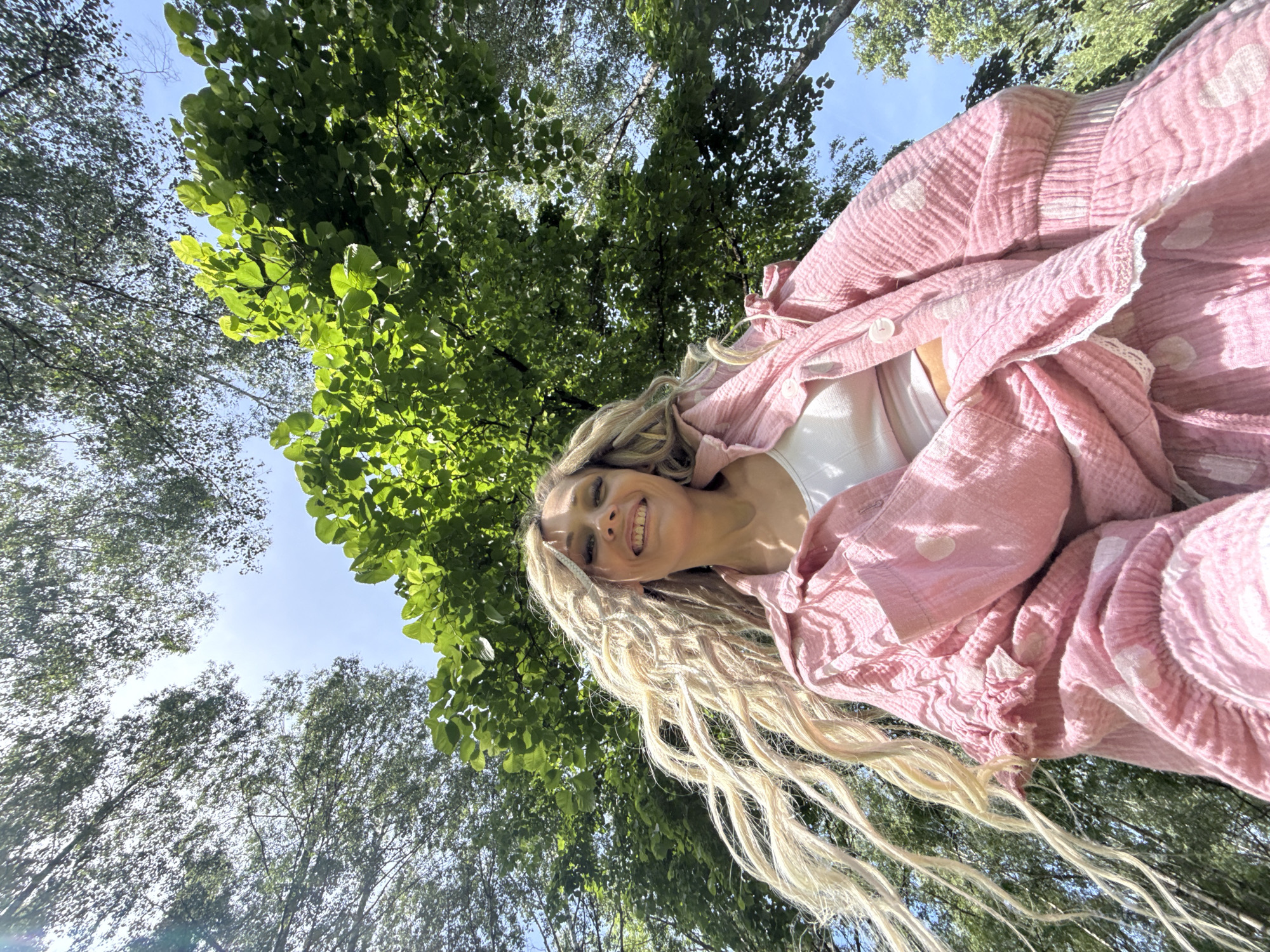 A low-angle upward photograph captures an adult female smiling directly at the camera in a heavily wooded outdoor setting in Rusanovo, Russia. The woman has long, light blonde hair with a distinct textured, wavy style, which appears wind-swept and flows down the sides of her face and shoulders. She is dressed in a loose-fitting, pink, crinkled gauze-like button-up shirt with white polka dots, worn over a white ribbed crop top. Matching pink bottoms, likely shorts or a skirt of similar fabric and pattern, are visible below. The background is dominated by a vibrant green tree canopy, with multiple shades of foliage filling the upper frame. Clear blue sky is visible through gaps in the leaves, suggesting bright daylight. A subtle lens flare is present in the upper left quadrant of the image.
