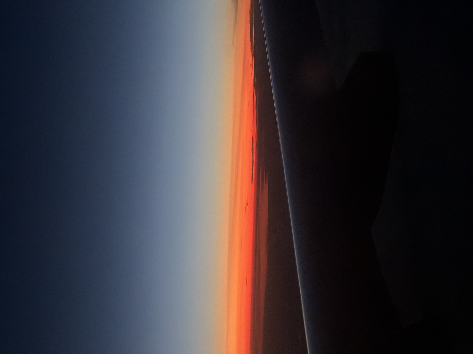 An aerial view captured from an airplane window shows a twilight sky over a dark landscape. The sky transitions from a deep blue at the top to vibrant orange and red hues near the horizon. The silhouette of an airplane wing occupies the foreground, its dark shape contrasting with the brightly colored sky. The land below is obscured by darkness, with faint lights suggesting a populated area, possibly the city of Perepechino, Russia, given the context. The scene conveys a sense of transition between day and night, as seen from high altitude.