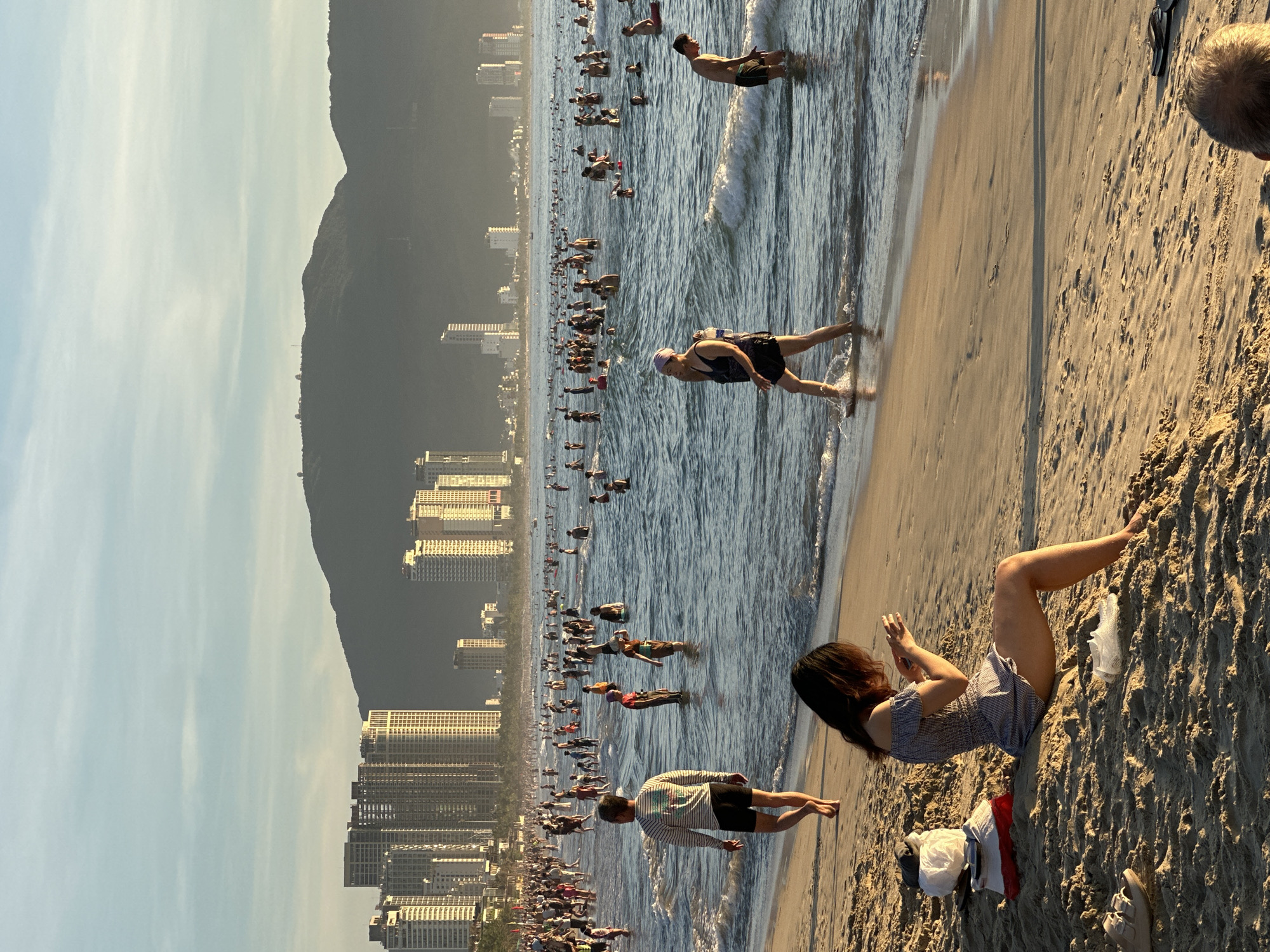 The image depicts a crowded beach scene in Đà Nẵng, Vietnam, with a backdrop of modern high-rise buildings and lush green mountains. The sun is low in the sky, casting long shadows and creating a warm, golden light across the sand and water. Numerous people are visible, many wading or swimming in the ocean, while others are walking along the shore or relaxing on the beach. In the foreground, a woman is seated on the sand, looking at her phone. To her left, a bag and some clothing items are placed on the sand, and a pair of sandals are visible in the lower left corner. Further down the beach, a man in a striped long-sleeved shirt and shorts walks away from the camera. The ocean is populated with many individuals, some in swimwear, engaged in various water activities. The city skyline dominates the background, with tall buildings reflecting the sunlight, and beyond them, the dark silhouette of mountains. The scene captures a lively and popular recreational area.