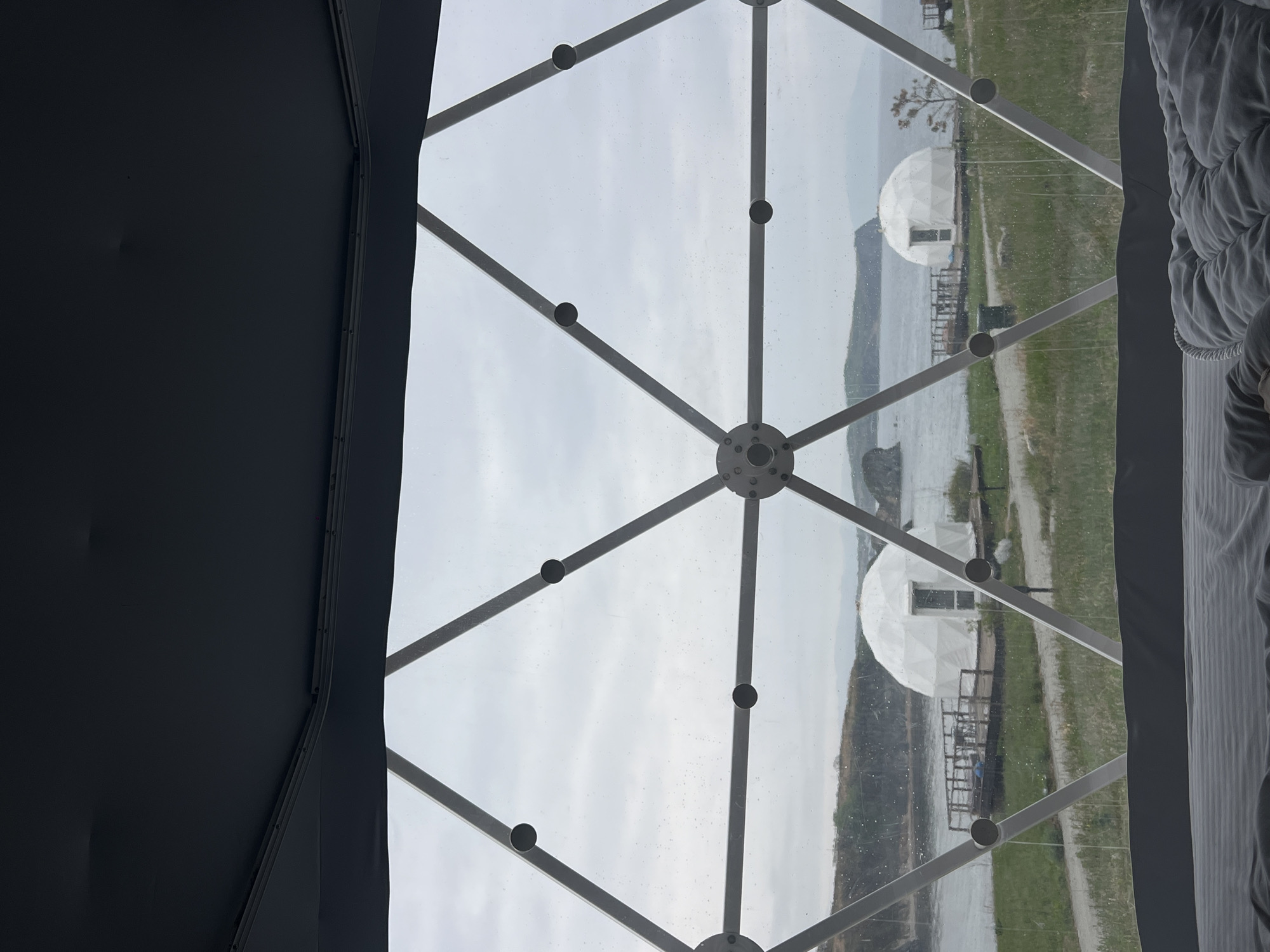 The image shows a view from inside a geodesic dome structure, likely a glamping site or similar accommodation. The framework of the dome is visible, creating a pattern of triangles and hexagons across the transparent material that forms the dome's outer shell. Through the dome, the outside scenery is visible, which appears to be a waterside area. Two dome-shaped structures are visible in the distance near the water, similar in construction to the one the photo is taken from. Between the buildings is a body of water with what looks like hills or a landform on the other side. To the right of the dome framework, there is the corner of a bed with white linens. The image is taken from inside the structure, looking outward. The photograph has been rotated and should be viewed 90 degrees counter-clockwise. Based on the prompt, the location is the city of Zarubino, Russia.