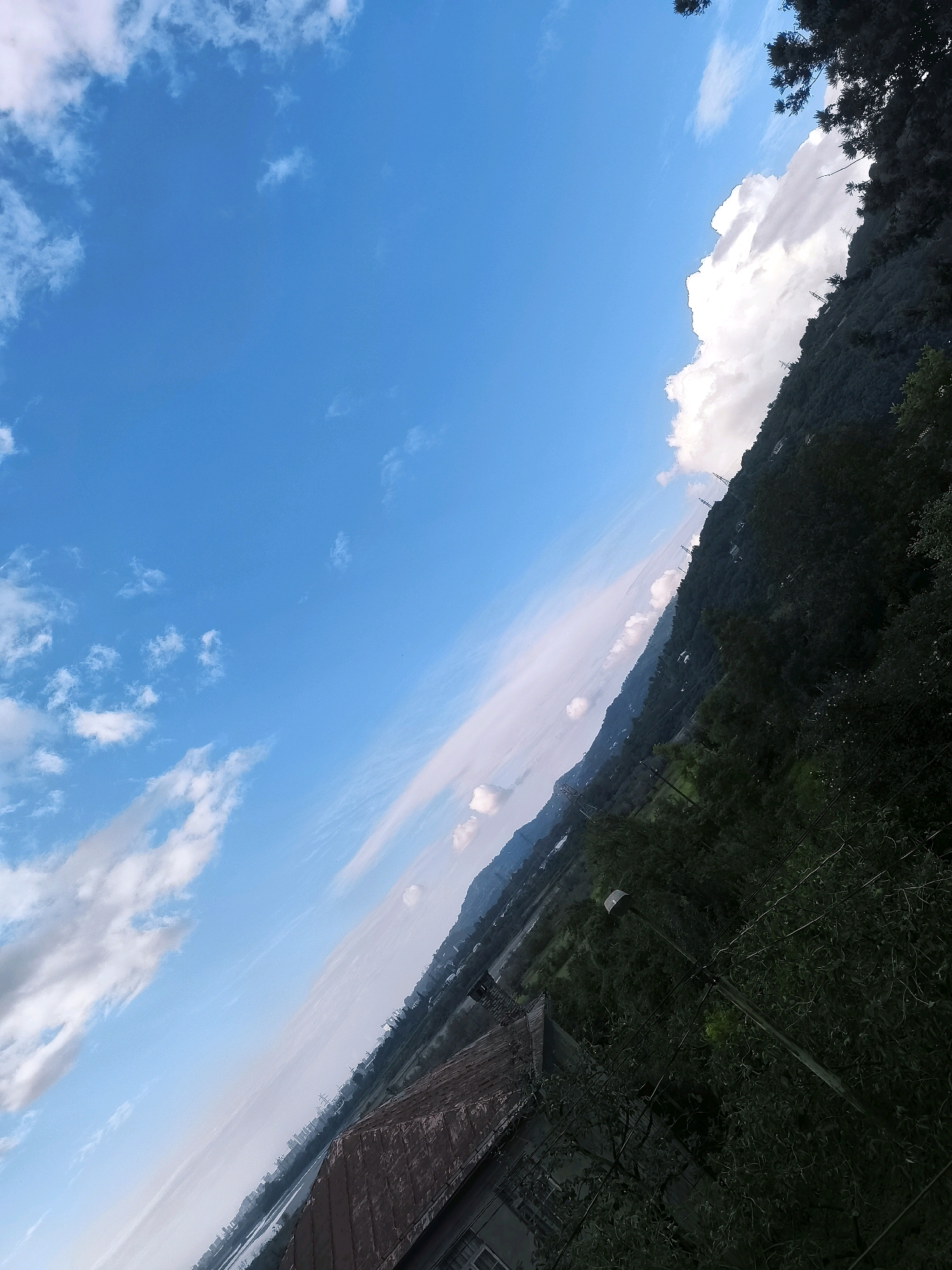 The image depicts a tilted landscape with a bright blue sky filled with scattered white clouds. In the foreground, the brown, textured roof of a building is visible, partially obscured by foliage. The middle ground shows a lush green hillside densely covered with trees, sloping upwards. Along the incline of the hill, several utility poles are visible. Further in the distance, a city skyline with various buildings and structures can be seen, bordering a body of water. The overall scene suggests an outdoor environment, likely on the outskirts of Batumi, Georgia, characterized by a blend of natural landscape and urban development under a clear sky.