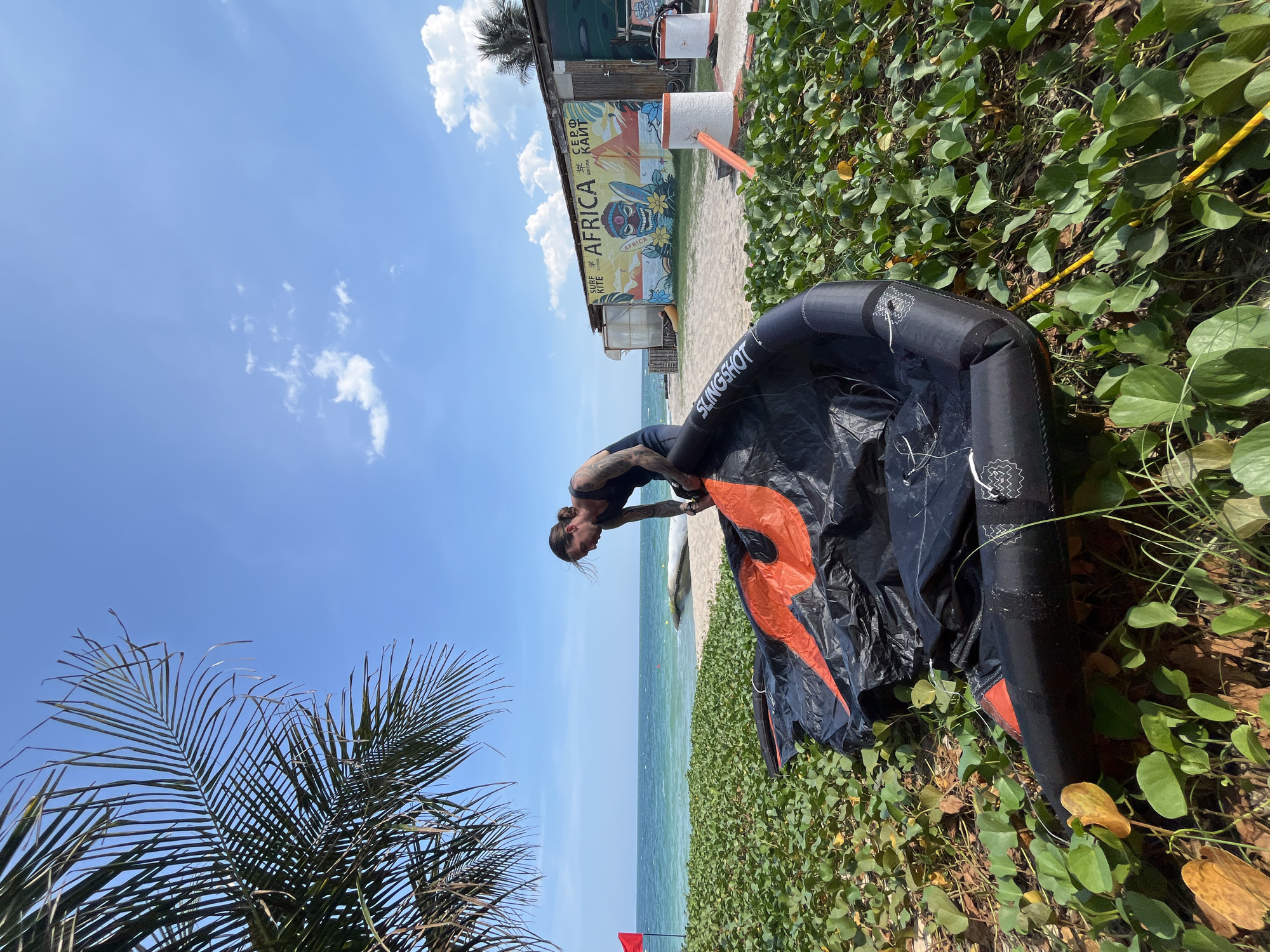 On a sunny day at the beach in Phu Thuy Ward, Vietnam, a man with tattoos on his arms and back is seen tending to a large, black and orange kite lying on the sand amongst lush green vegetation. He is wearing a dark tank top and appears to be adjusting the kite. In the background, the turquoise ocean meets a clear blue sky with a few scattered clouds. A red flag flies on a pole to the left, and a building with a colorful mural depicting "AFRICA SURF KITE SCHOOL" stands further down the beach. The scene captures a moment of preparation for watersports on a tropical coast.
