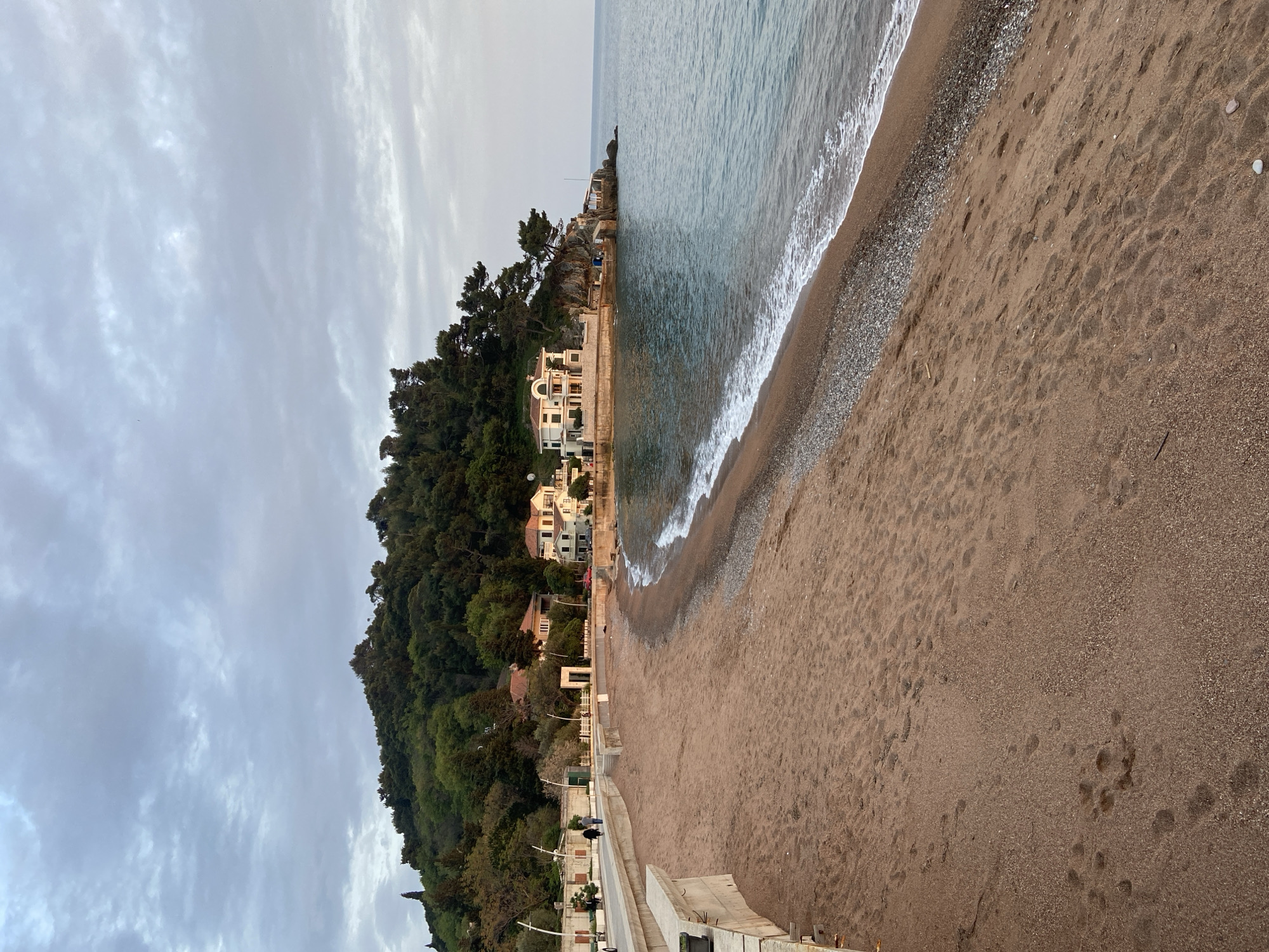 This image depicts a coastal scene in Petrovac centar, Montenegro, under an overcast sky. The foreground features a wide, brownish-tan sandy beach marked with numerous footprints, transitioning to a strip of small pebbles where the gentle waves of the sea meet the shore. A concrete promenade runs along the left edge of the beach, with some distant figures faintly visible walking along it.
To the left of the promenade, a steep, lush green hillside covered in dense trees dominates the landscape. Several buildings, characterized by their white or light-colored facades and traditional architecture, are nestled among the trees along the coastline and further up the hill. One prominent building with a distinctive archway is visible closer to the beach.
The sea occupies the right portion of the frame, displaying calm blue-grey waters with white foam from small breaking waves. The horizon line is indistinct due to the cloudy conditions. The sky above is entirely covered with light grey, streaky clouds, indicating an overcast day.
No specific activities are clearly taking place, and no discernible text is visible on any signs or structures. The scene conveys a tranquil atmosphere in a seaside town.