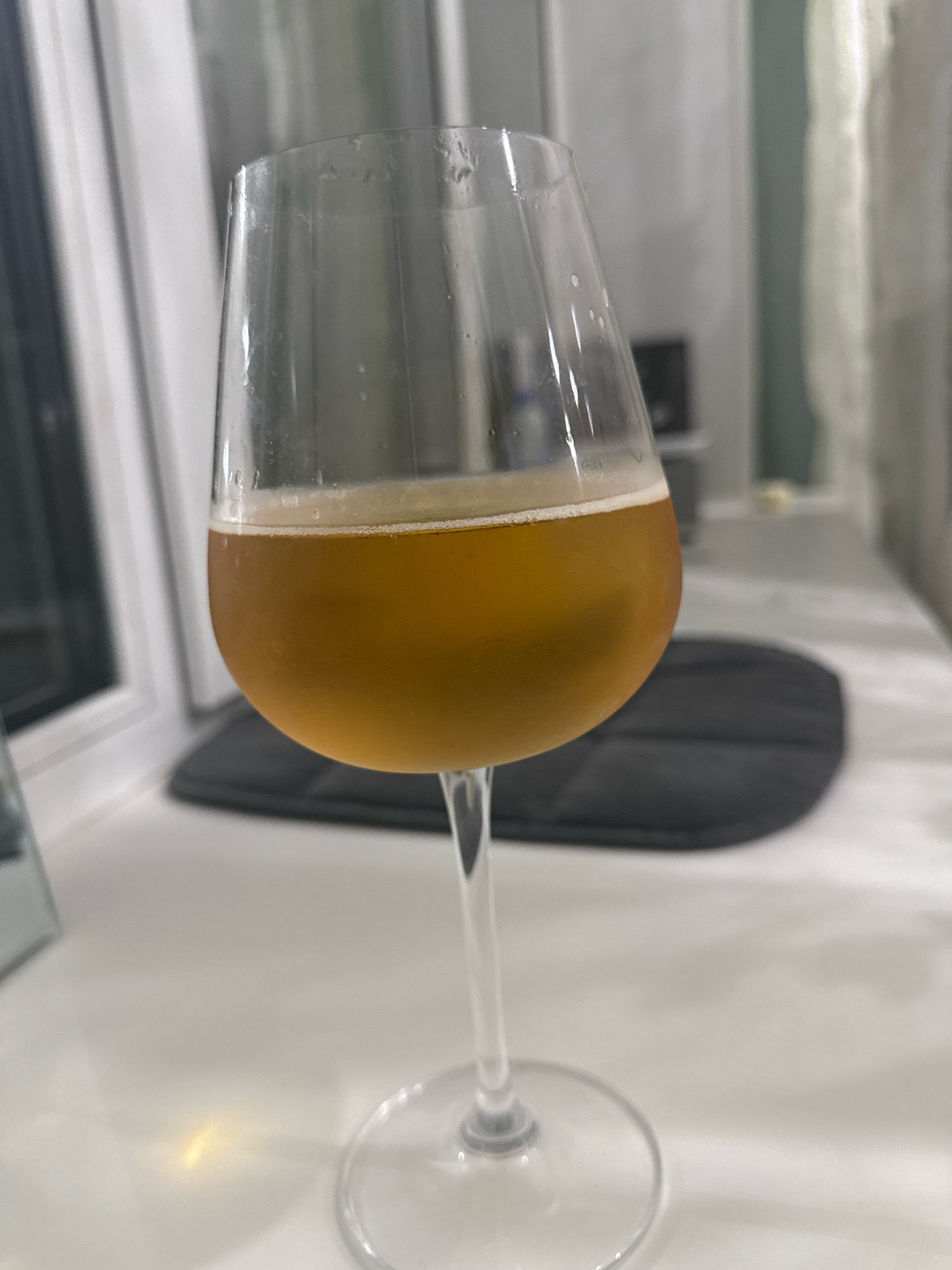 A close-up, eye-level shot shows a stemmed wine glass filled with a golden-amber colored liquid and a thin layer of foam at the top. The glass is clear and appears to be made of glass, with condensation visible on the exterior. The glass is placed on a white, flat surface that reflects a yellowish light. In the background, slightly out of focus, is a dark, oval-shaped mat or coaster. To the left of the glass, a white window frame is visible, with vertical blinds or curtains behind it. The overall environment suggests an indoor setting, likely a home or a bar, in Cheboksary, Russia. No people or animals are present in the frame, and no specific actions are depicted, other than the static placement of the glass.