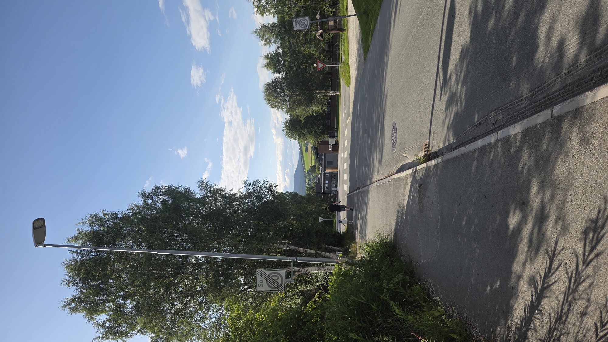 The image depicts a paved road and sidewalk in Tynset, Norway, on a sunny day with a blue sky and scattered clouds. The scene is lined with lush green trees on the left and right, with a backdrop of rolling hills and buildings.
On the sidewalk to the left, a person, likely an adult, is walking with a child. The adult is wearing dark shorts and a dark t-shirt, and the child is wearing a light-colored hat and possibly a light-colored top. They are walking away from the camera, towards an intersection in the distance.
Several signs are visible. On the left side of the road, a speed limit sign is mounted on a pole, indicating "SONE 50", suggesting a 50 km/h speed limit zone. Another sign, a yield sign (inverted triangle), is visible near the intersection. On the right side of the road, a similar "SONE 50" sign is present, along with a wooden post or small shelter.
The road surface shows tire tracks, and a manhole cover is visible on the pavement. Tree shadows are cast across the sidewalk and road, indicating the direction of the sun. In the distance, several buildings, including a multi-story structure with visible windows, are situated. The overall environment suggests a semi-urban or suburban area with a mix of residential and commercial buildings, surrounded by natural greenery.