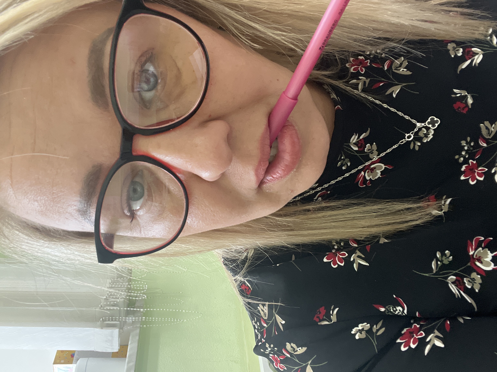 A woman is holding a pink lip liner pencil to her lips while wearing black and red glasses. She has light blonde hair and is wearing a black dress with a red and white floral pattern. A silver necklace with a clover pendant is visible. The background shows a green wall and a window with white blinds. The image location is Chaikovsky, Russia.