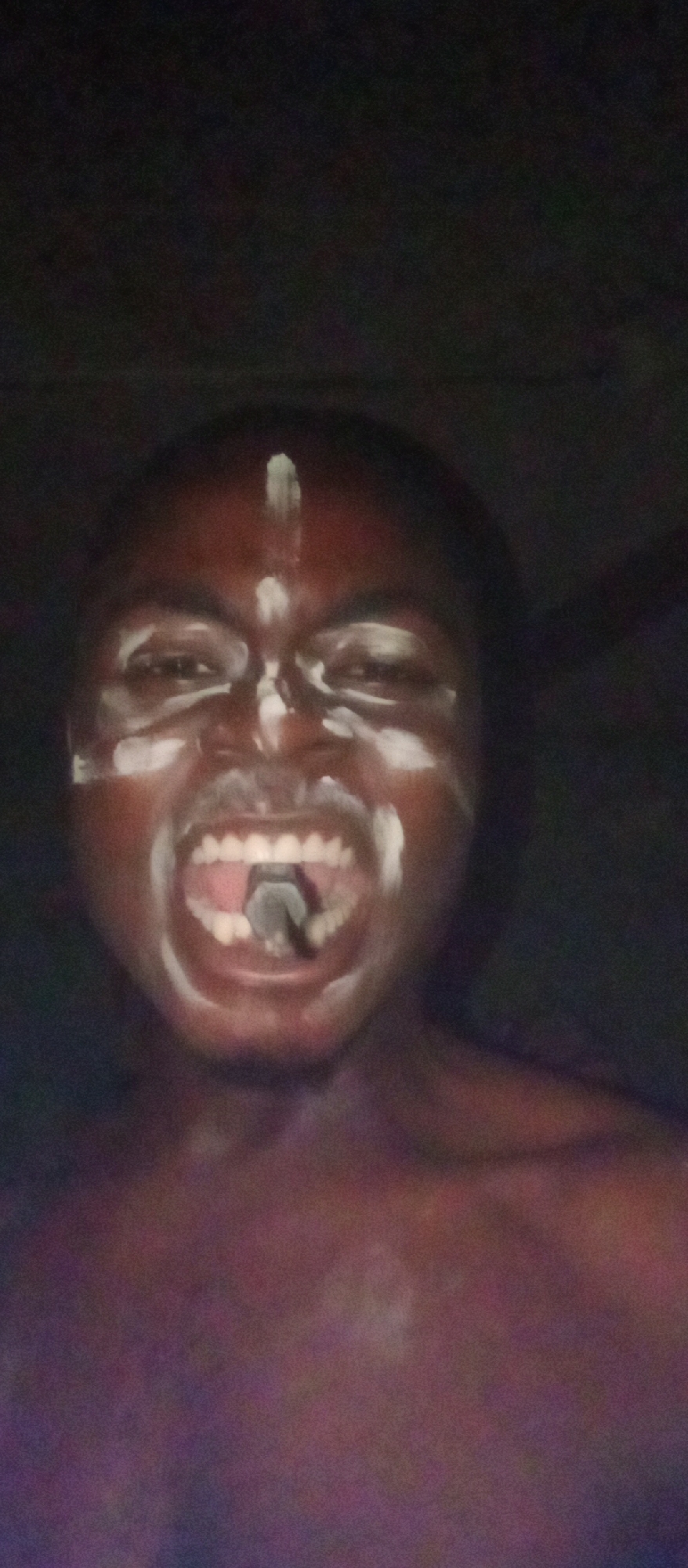 A close-up image shows a person, likely male, with dark skin, in what appears to be a dimly lit or dark environment. The individual's face is painted with distinct white markings: a vertical line runs from the forehead down to the nose, and smears of white paint encircle the eyes, mark the cheeks, and extend across the chin. The person's mouth is wide open in what suggests a shout, scream, or vocalization, revealing their teeth. A dark, cylindrical object or scroll is visible inside the mouth, resting on the tongue. The upper body appears bare, or partially covered by dark clothing that blends with the background. The background is uniformly dark and indistinct, providing no specific location cues. The setting appears to be at night or in a very poorly lit indoor space. There is no visible text in the image. The context for this image is Ogidi, Nigeria.