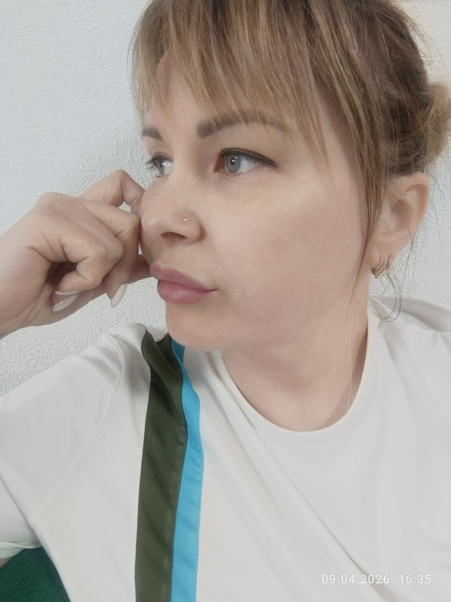 A fair-skinned woman with light brown hair pulled back into a bun, featuring bangs, is captured in a close-up, side-profile shot. She has light-colored eyes, a nose piercing, and wears hoop earrings. She rests her fingers on her cheek with a thoughtful expression. She's wearing a white shirt with a broad, dark green stripe next to a light blue stripe down one side. The background is a plain, textured white wall. The timestamp "09.04.2026 16:35" is visible in the lower right corner of the image. No further information is available from the image to determine its location in Chaikovsky, Russia.