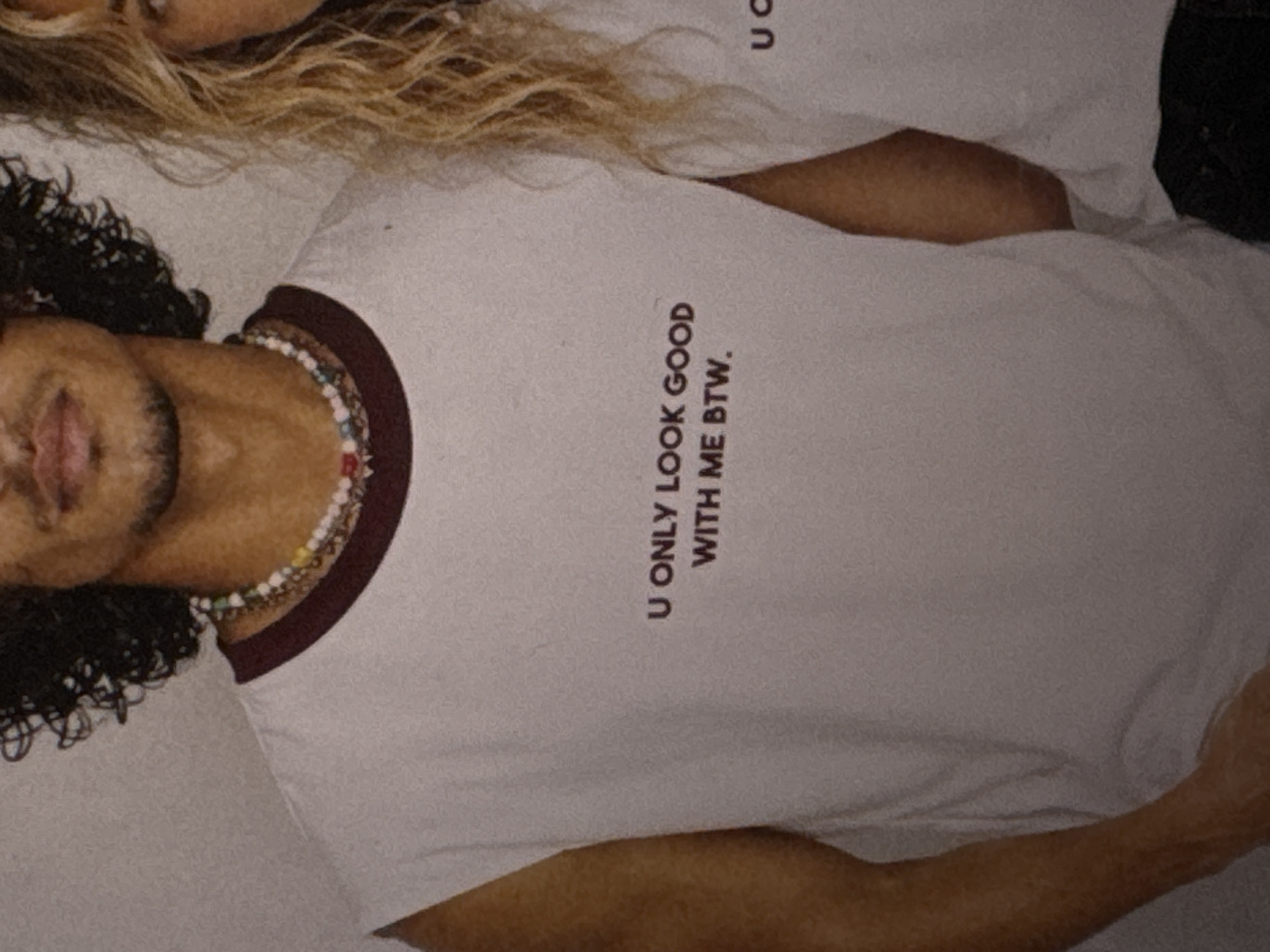 Two individuals, a man and a woman, are positioned close together. The man, in the foreground, has dark, curly hair and is wearing a white t-shirt with a maroon trim around the collar. The t-shirt has text printed vertically on it. The woman, in the background, has blonde, wavy hair. The scene appears to be an indoor setting, possibly a studio due to the soft lighting and plain background. No specific time of day, weather, or emotional cues are discernible.
The text on the man's t-shirt reads: "U ONLY LOOK GOOD WITH ME BTW."