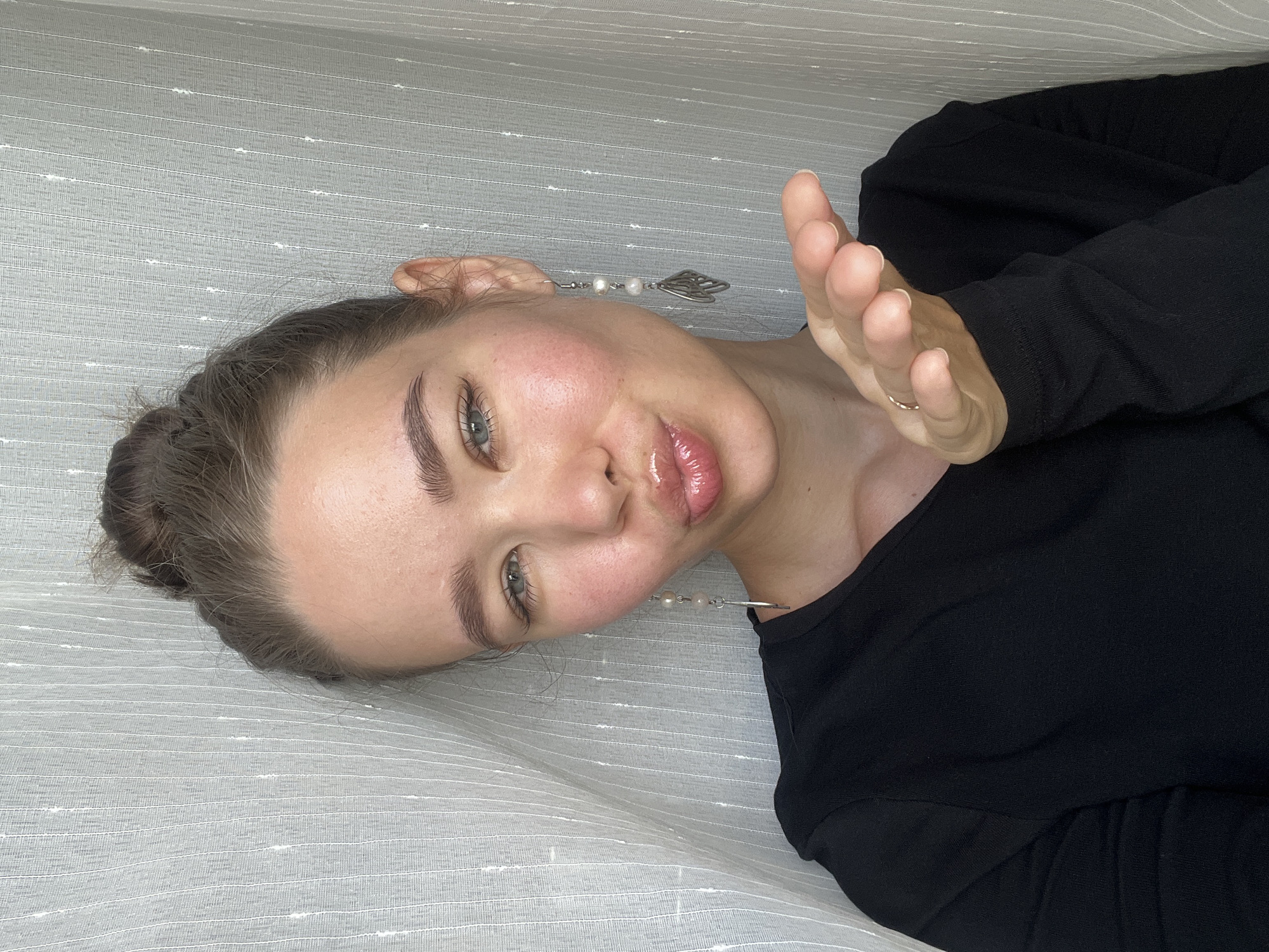 A person with light skin and green eyes is lying on a textured white surface. Their dark hair is pulled back in a bun. The person is wearing a black top. They have a silver earring in their right ear and a gold ring on their left hand. The person's right hand is raised with the fingers partially extended. The image does not provide information about the location or specific actions.