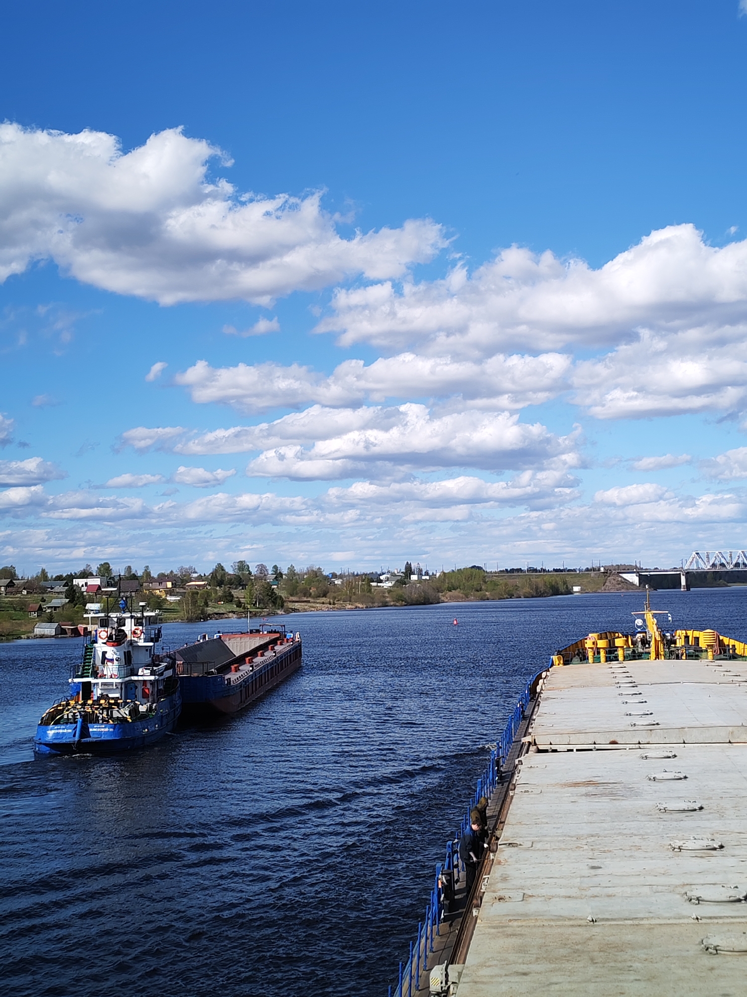 The image shows a wide river under a partly cloudy blue sky. In the foreground is a large, flat, light-gray barge, with blue railings and some yellow equipment on its deck. Two people are visible on the edge of the barge near the railing. To the left, a blue tugboat is attached to a dark-colored barge loaded with material. Both vessels are moving along the river, creating wakes in the water. On the far bank, there is a town or village with buildings and trees. A bridge is visible in the distance. This appears to be the location near Kochino, Russia.