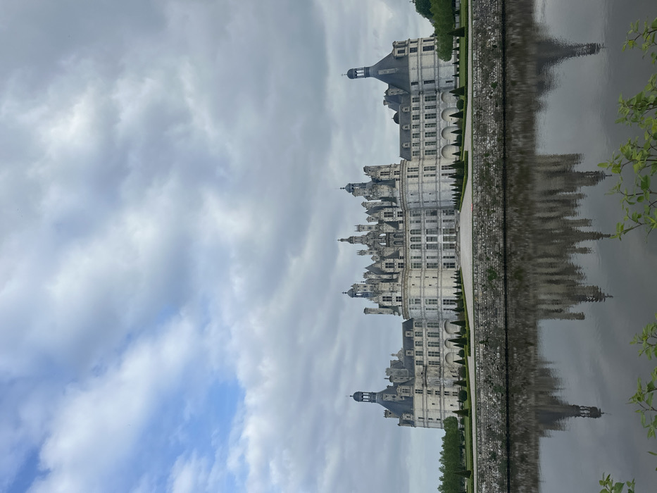 The Château de Chambord, located in the Loire Valley of France (latitude: 47.617, longitude: 1.515), is pictured from a distance in front of a wide moat. The large white castle is made of stone, with a central tower and numerous smaller towers and spires. The castle is symmetrical, with two identical wings extending out from the center. The castle is surrounded by a moat, which is reflected in the water. The water appears calm and still, and the reflection of the castle is clear. The sky is cloudy and overcast. There are no people visible in the picture. A few trees are visible in the foreground and on the banks of the moat, and there are a few trees visible in the background behind the castle.