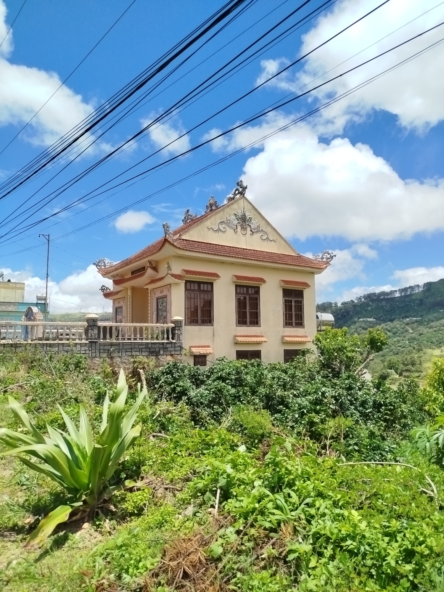 A medium shot captures a light yellow building with a red-tiled roof and decorative eaves, resembling a traditional Vietnamese or Chinese architectural style, situated on a slight incline. The building has multiple windows with dark brown frames and is fronted by a low stone and concrete wall with a decorative railing. Lush green vegetation, including a large leafy plant in the foreground, surrounds the structure and the hillside behind it. The sky above is bright blue with scattered white clouds, and several black power lines are visible stretching across the top of the frame. In the distance, a treed hillside is visible. A single person, a woman wearing a light blue top and dark pants, stands near the railing on the left side of the building, facing towards the left, partially obscured by vegetation. The scene appears to be located in a rural or semi-rural area, possibly on the outskirts of Trường An city, Vietnam, given the architectural style and natural landscape.