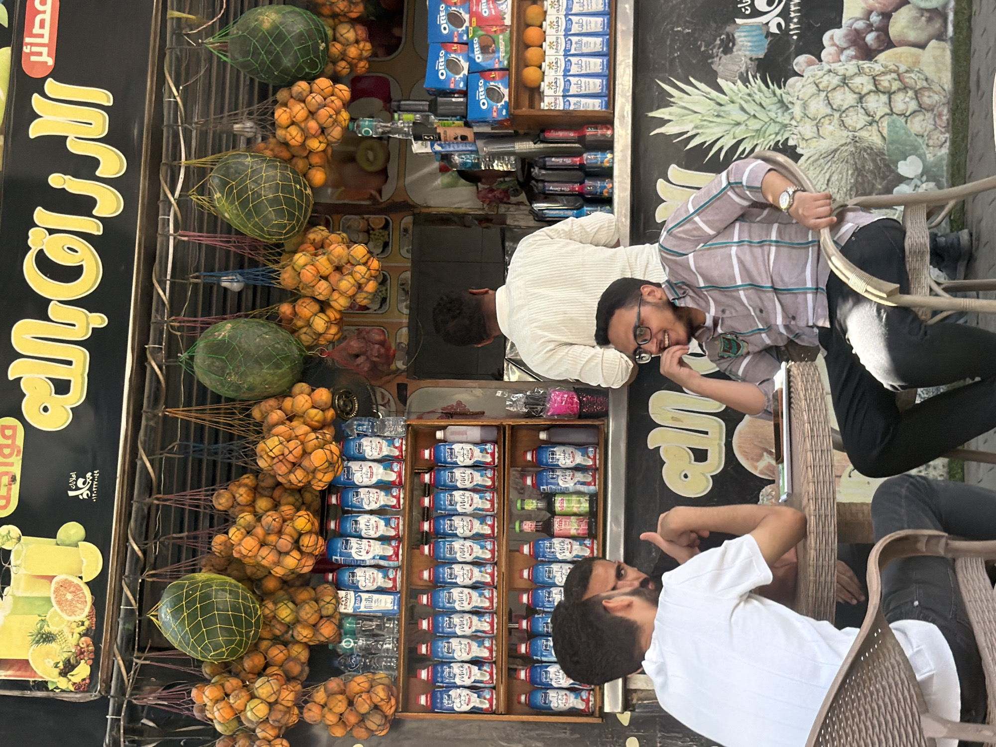 The image depicts an outdoor juice stand in Cairo, Egypt. A brightly colored sign with Arabic text hangs above, advertising various juices. Below the sign, an assortment of fresh fruits, including oranges and large green watermelons, are displayed, some hanging in netting. Shelves are stocked with numerous bottles of milk and juice, as well as boxes of Oreo cookies.
In the foreground, three men are seated at a circular table. Two of the men are looking towards the juice stand, while the third man, wearing glasses and a checkered shirt, is looking to his right and smiling. The environment appears to be a busy street, with the juice stand serving as a focal point.