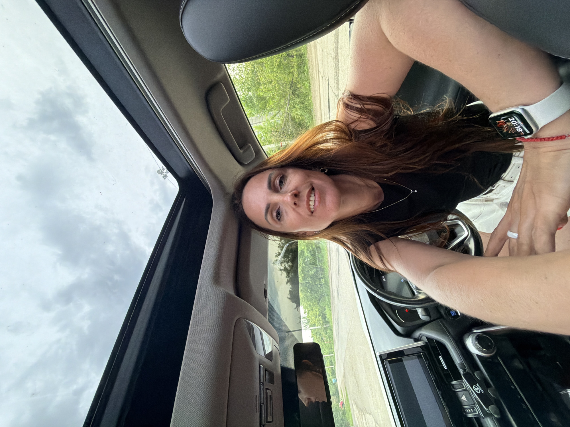 A woman with long brown hair is sitting in the driver's seat of a car, looking at the camera and smiling. She is wearing a black top and white shorts. The car has a sunroof open, and the sky is visible. The interior of the car is visible, including the dashboard, steering wheel, and a touchscreen. The woman is wearing a white smartwatch on her left wrist. The car appears to be parked on a road with trees and vegetation in the background. There is no indication of the city of Birobidzhan, Russia, in the image.