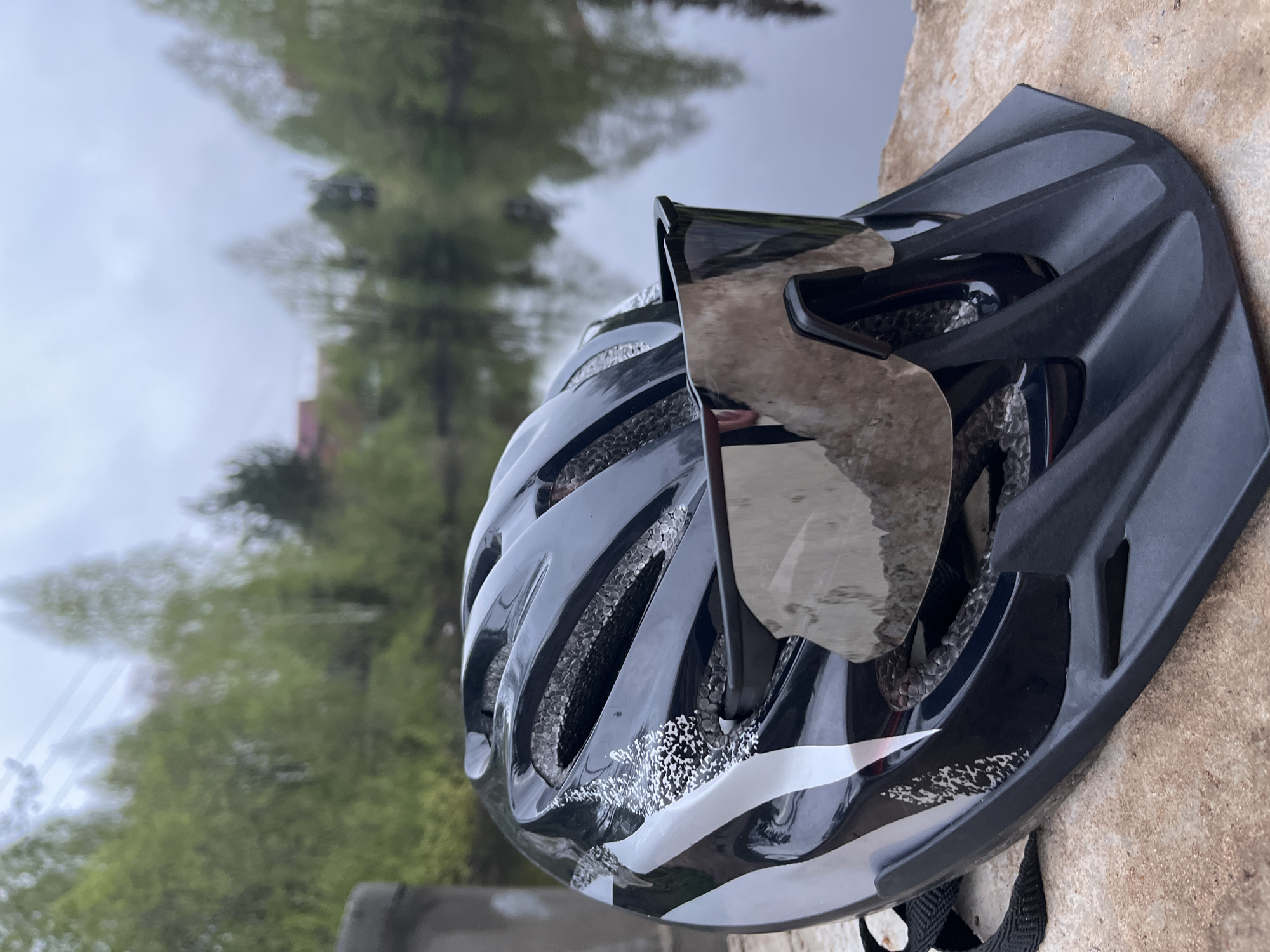 A black and white bicycle helmet rests on a concrete surface. The environment in the background includes trees and sky, reflected in what appears to be a body of water. While specific location data is unavailable, it is assumed to be outdoors and close to a natural environment. The location is assumed to be the city of Chaikovsky, Russia, as mentioned. No people or interactions are directly visible in the image.