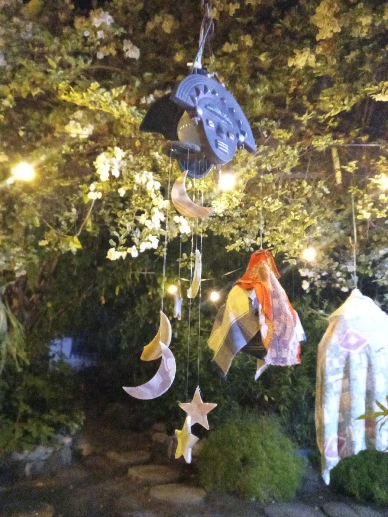 A medium shot shows an outdoor scene at night, possibly in El Limón, Venezuela, with a wind chime hanging prominently in the foreground. The wind chime is composed of a dark-colored, possibly metallic, top component with crescent moon and star-shaped pendants suspended below. Adjacent to the wind chime, decorative fabrics or cloths are also hanging, one with an orange and multi-colored pattern and another with a lighter-colored print.
The background is filled with dense green foliage and flowering plants, illuminated by string lights that suggest a garden or patio setting. The overall scene is dimly lit, creating a soft and somewhat obscured view of the surroundings. The image captures a moment of tranquility and artistic expression in a natural outdoor environment.