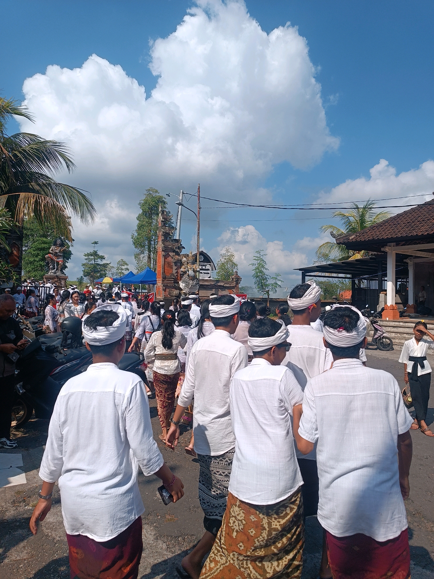 A group of people are walking down a street in the city of Tabanan, Indonesia. Many of the individuals are wearing traditional white shirts and sarongs, with some also wearing white headbands. The street is lined with buildings, including a temple with ornate carvings. There are also trees, power lines, and a blue sky with white clouds overhead. Several motorcycles are parked on the side of the street. The people appear to be participating in a procession or event.