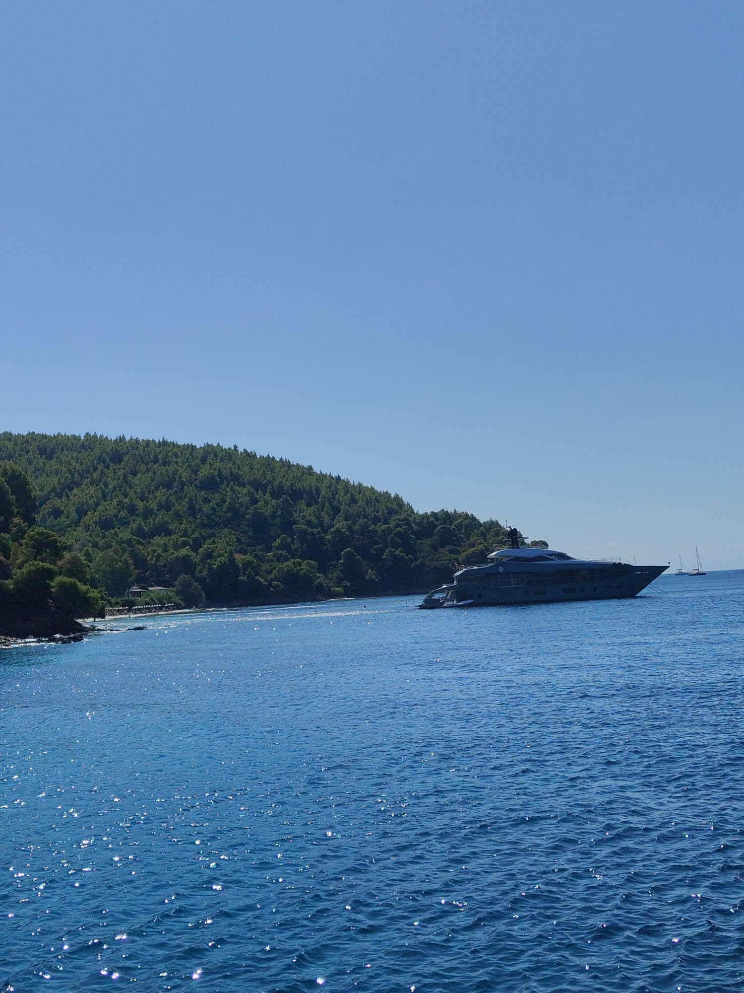 A wide-angle, eye-level shot captures a serene coastal scene. The sky is a clear, cloudless blue, taking up the top third of the frame. Below, the turquoise sea shimmers with sunlight, its surface rippled by a gentle breeze. In the middle ground, a large, dark-grey yacht is anchored offshore. The yacht is sleek and modern, with multiple decks and a pointed bow. To the left of the yacht, a lush green hillside covered in dense trees slopes down towards the water. A small stretch of beach is visible at the base of the hill, with a few beach umbrellas and loungers. The scene suggests a tranquil summer day in a Mediterranean location, likely Greece, given the mountainous terrain and the style of the yacht.