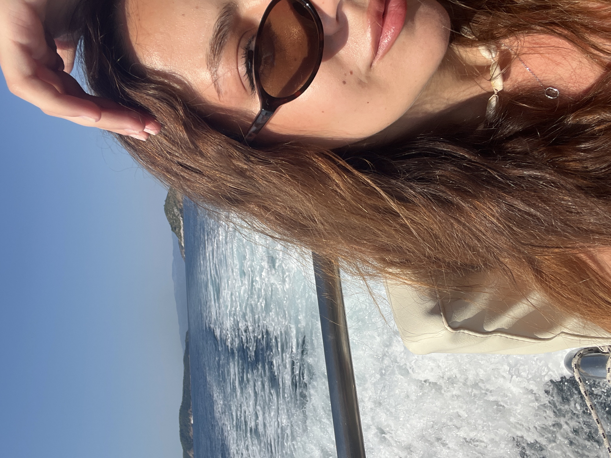 A young woman with long brown hair and sunglasses poses for a selfie on a boat sailing in the sea. The boat is leaving a white wake behind it. In the background, there are green islands and a clear blue sky. The woman is wearing a light-colored jacket and a shell necklace. The sunlight is bright, casting shadows on her face. The scene appears to be in a coastal area, possibly near the island of Skiathos, Greece, given the presence of islands and the clear blue waters typical of the Aegean Sea.