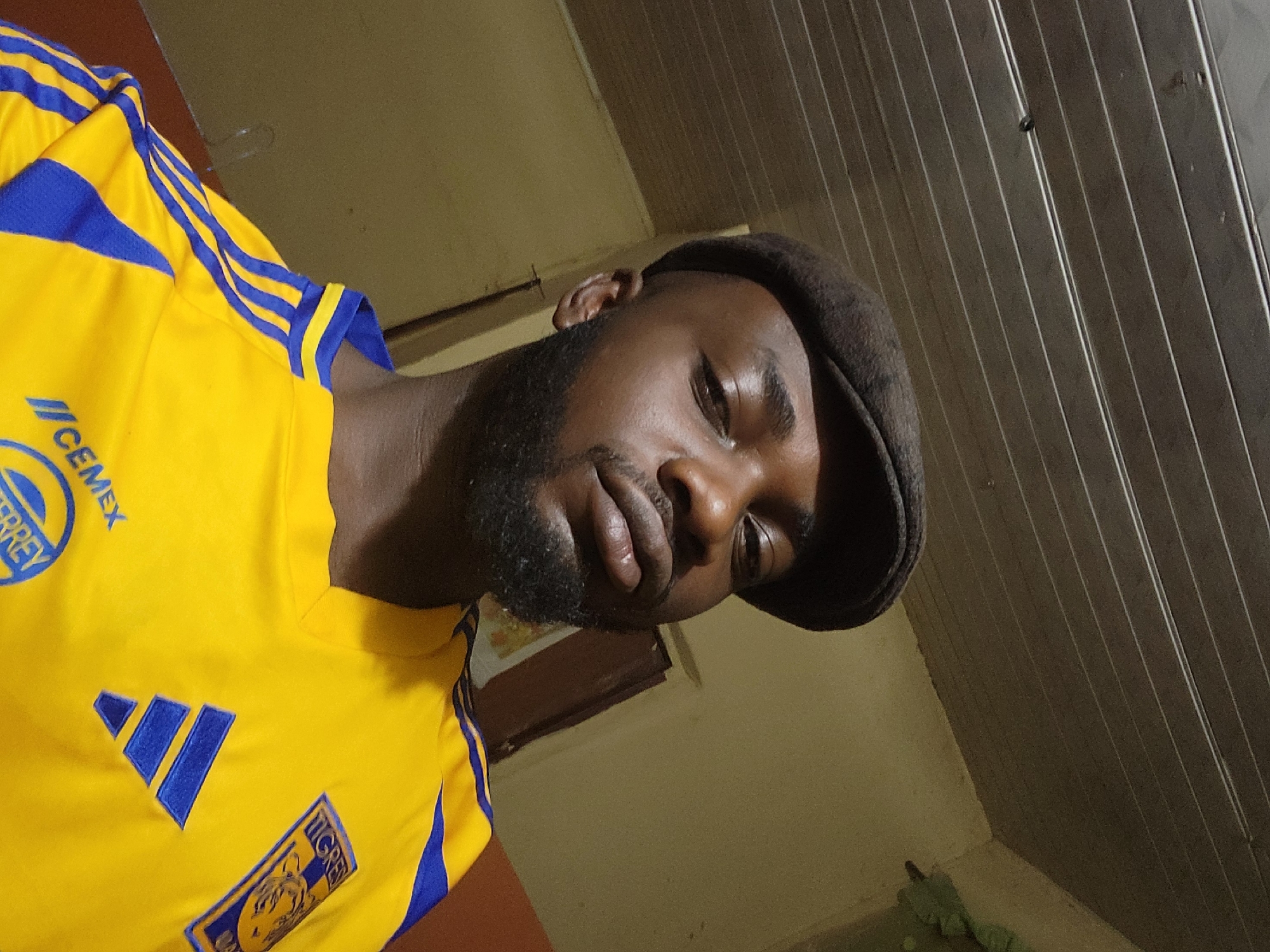 A portrait-style, low-angle, selfie shows a Black man wearing a flat cap and a yellow and blue striped jersey. He has a beard and is looking directly at the camera with a neutral expression. The background is dimly lit and shows a section of what appears to be a ceiling with horizontal planks, and a wall painted in a yellowish hue. The man is wearing a yellow jersey with blue Adidas stripes on the sleeves and an emblem on the right chest that appears to be a sports team logo, possibly Tigres UANL. The bottom of the jersey has "CEMEX" and "MONTERREY" written on it. There is a faint shadow on the left side of the man's face, suggesting the light source is to his right. The overall impression is of an indoor setting, possibly a bedroom or living area. The request for location in Jos, Nigeria cannot be fulfilled based on the visual information provided in the image.