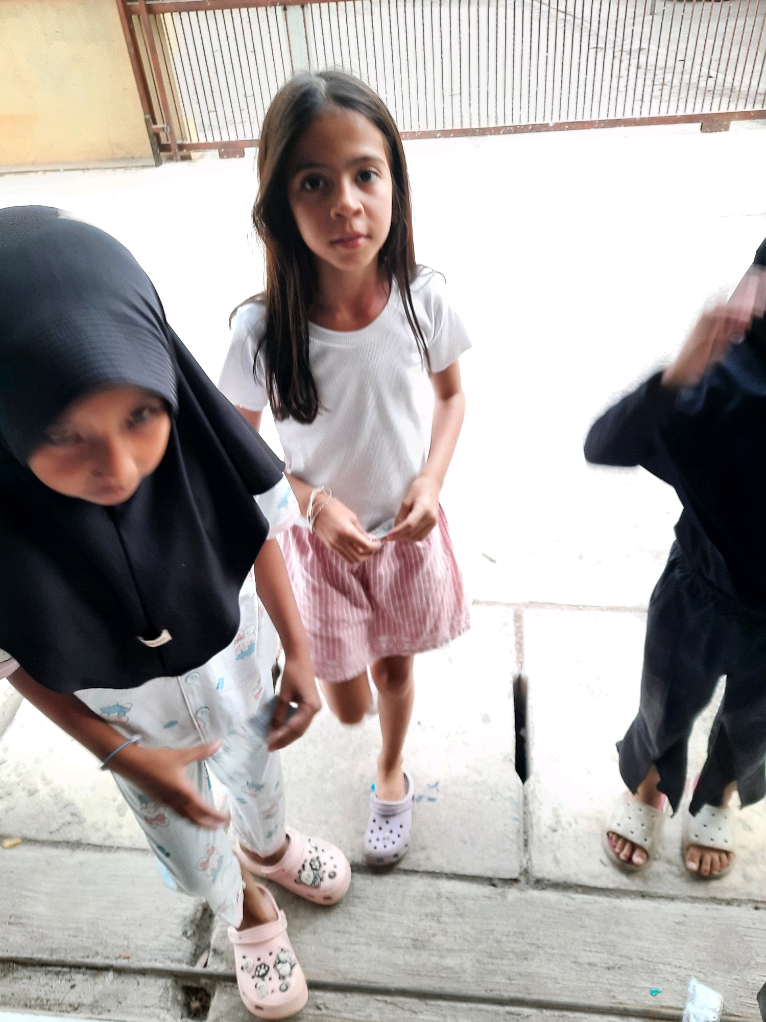 Three young girls are standing in a narrow outdoor alley or walkway. The girl in the center is facing forward, looking at the camera, and is wearing a white t-shirt and a pink and white striped skirt. To her left, a girl wearing a black hijab and patterned pajama pants is looking down, with her face partially obscured. To the right of the center girl, another person is partially visible, wearing a black shirt and pants, with their right arm blurred as if in motion. All three girls are wearing clogs on their feet. The background consists of a metal grate fence above a pale wall, and the ground is a concrete walkway with drainage grates. The lighting suggests it is daytime.