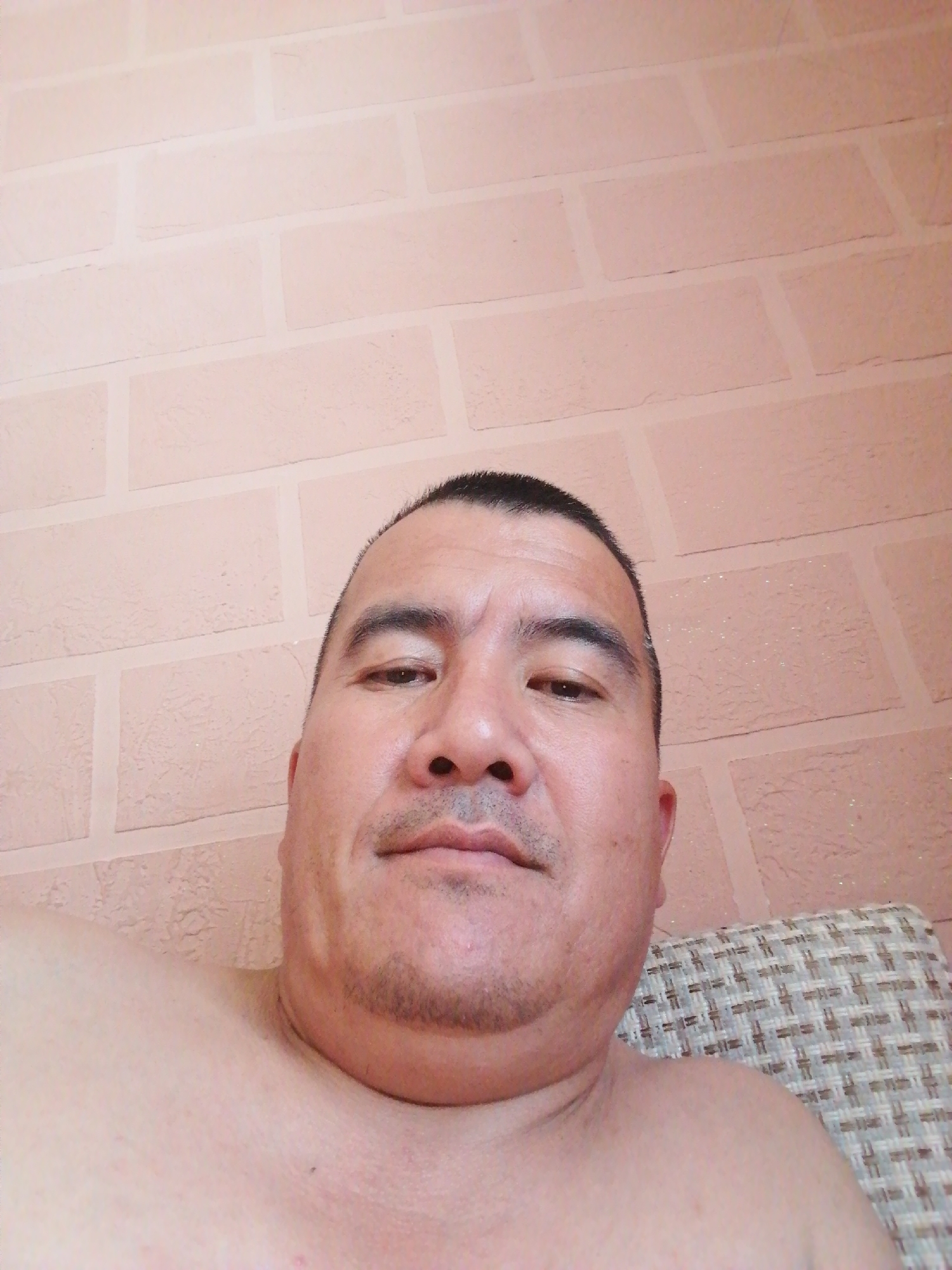 The image shows a close-up, overhead shot of a man's face and upper chest. He is looking directly at the camera. The man has dark hair, a mustache, and a light beard. His skin appears to be fair. He is shirtless, and his shoulders and upper chest are visible. Behind him, a textured, pale pink brick wall is visible. To the right, a portion of a woven chair or cushion is visible. The lighting is somewhat dim, with some specular highlights on the wall. This scene is not identifiable as being in Gumbaz, Uzbekistan.